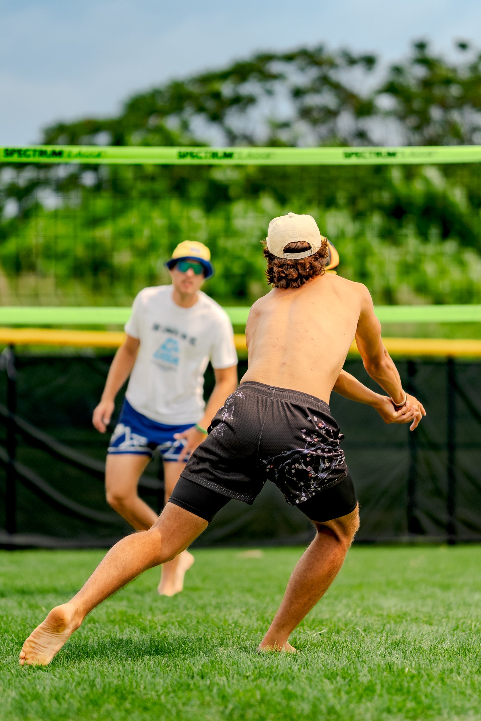 A player in black shorts with a floral pattern prepares to hit a volleyball, with another player in a white shirt and blue shorts in the background.