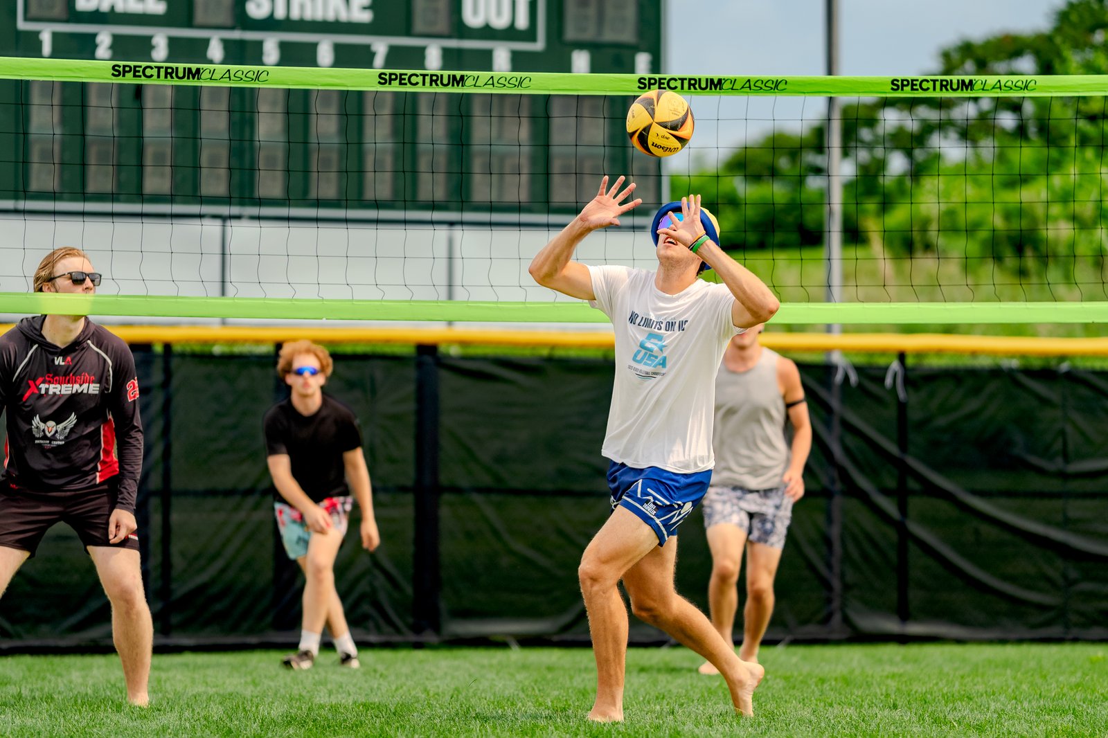 A player in a white shirt and blue shorts attempts to dig a volleyball near the net, with other players in black and red and black tops visible.