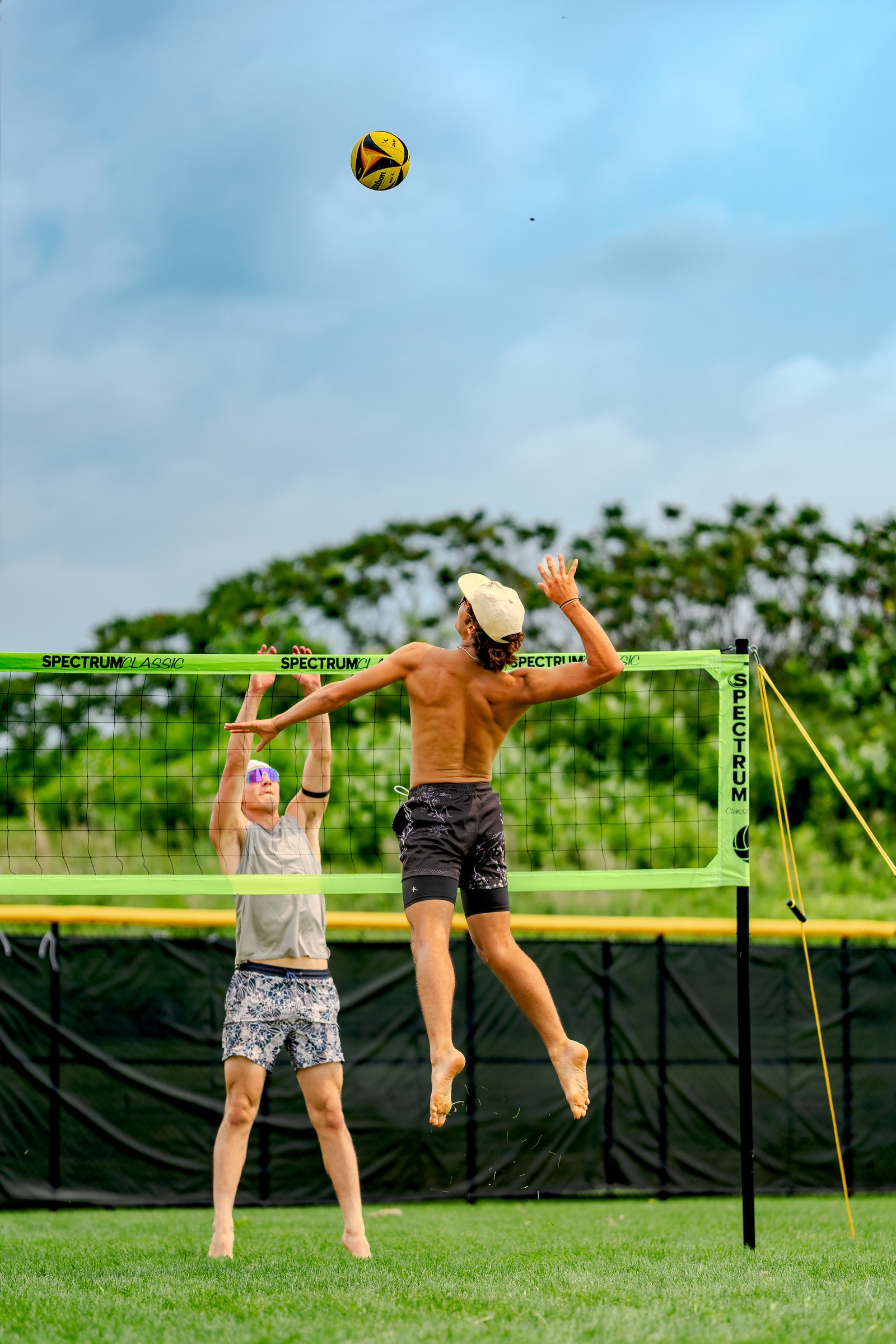Two players jump near the net as a volleyball flies overhead, one player in a grey top and patterned shorts, the other in black patterned shorts.