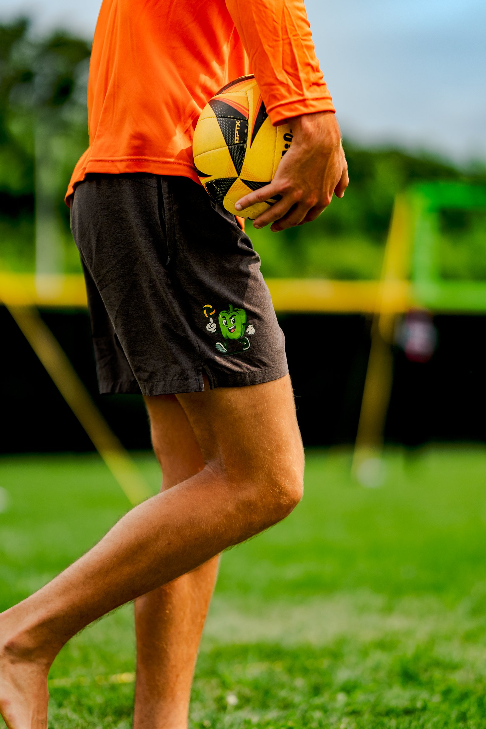 A player in an orange shirt and dark shorts holds a volleyball on a grassy court with a yellow net in the background.