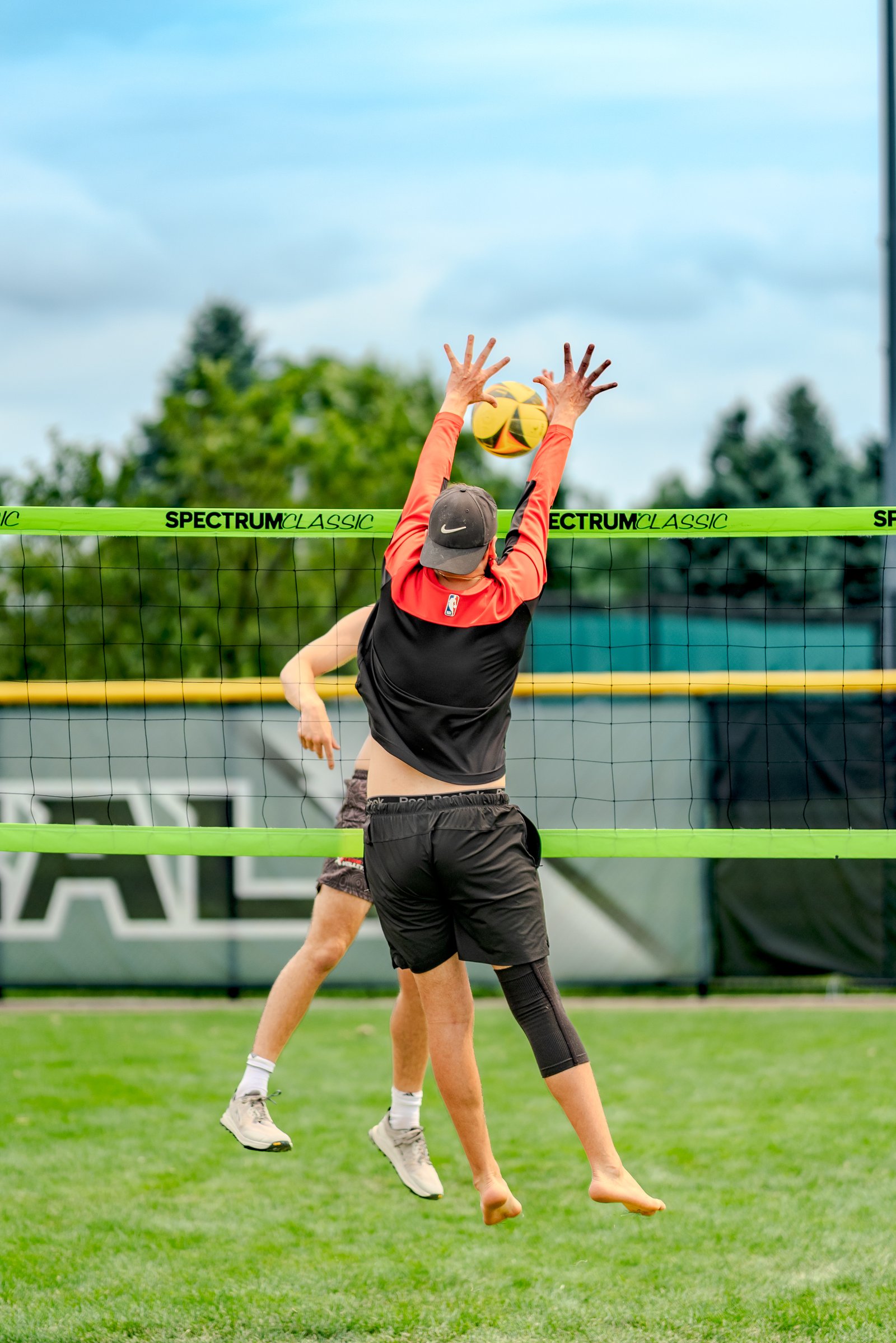 A player in a black and orange top jumps to block a volleyball near the net, with another player in black shorts visible behind.