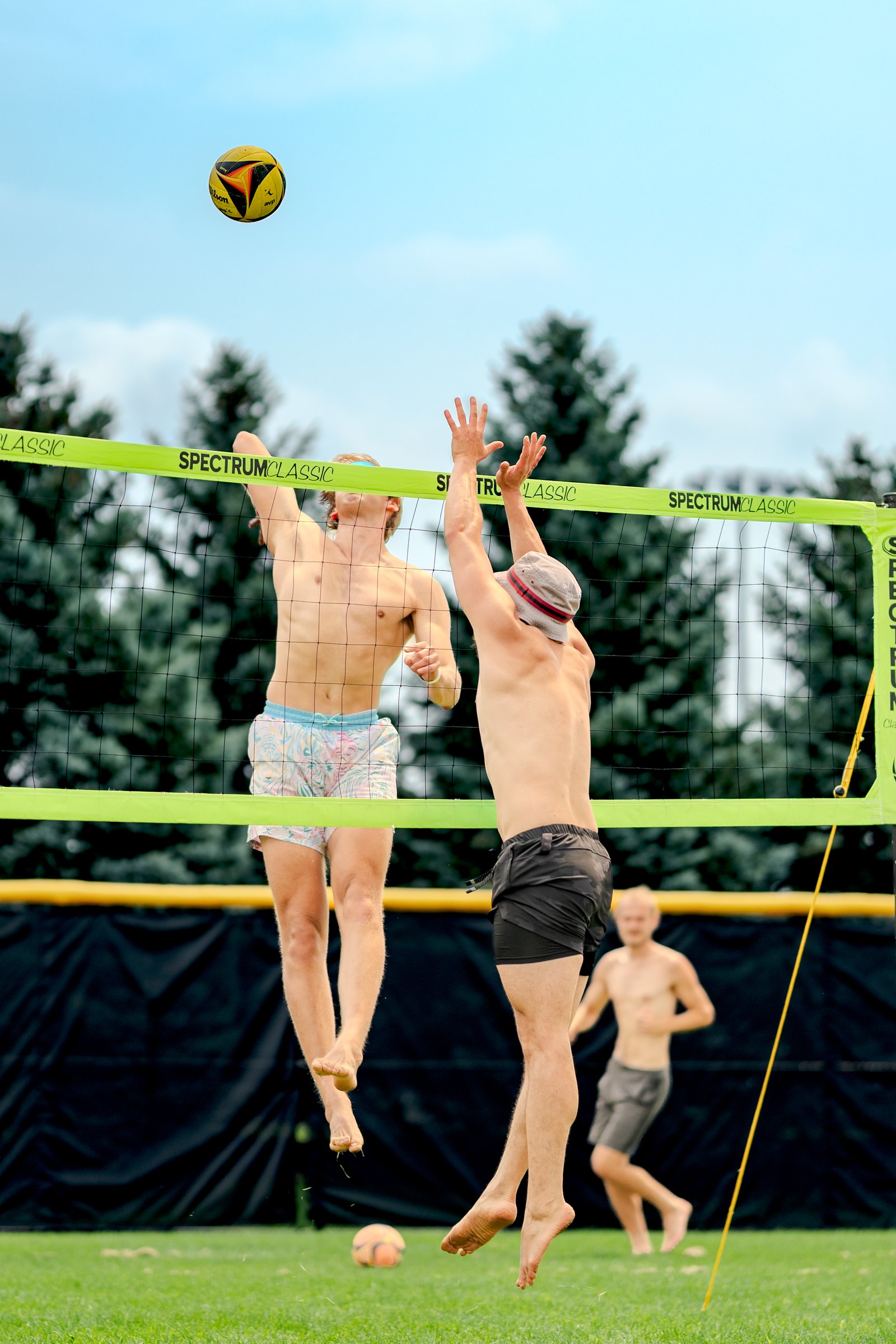 A player in patterned shorts jumps to hit a volleyball while a player in black shorts reaches to block, with another player in gray shorts in the background.