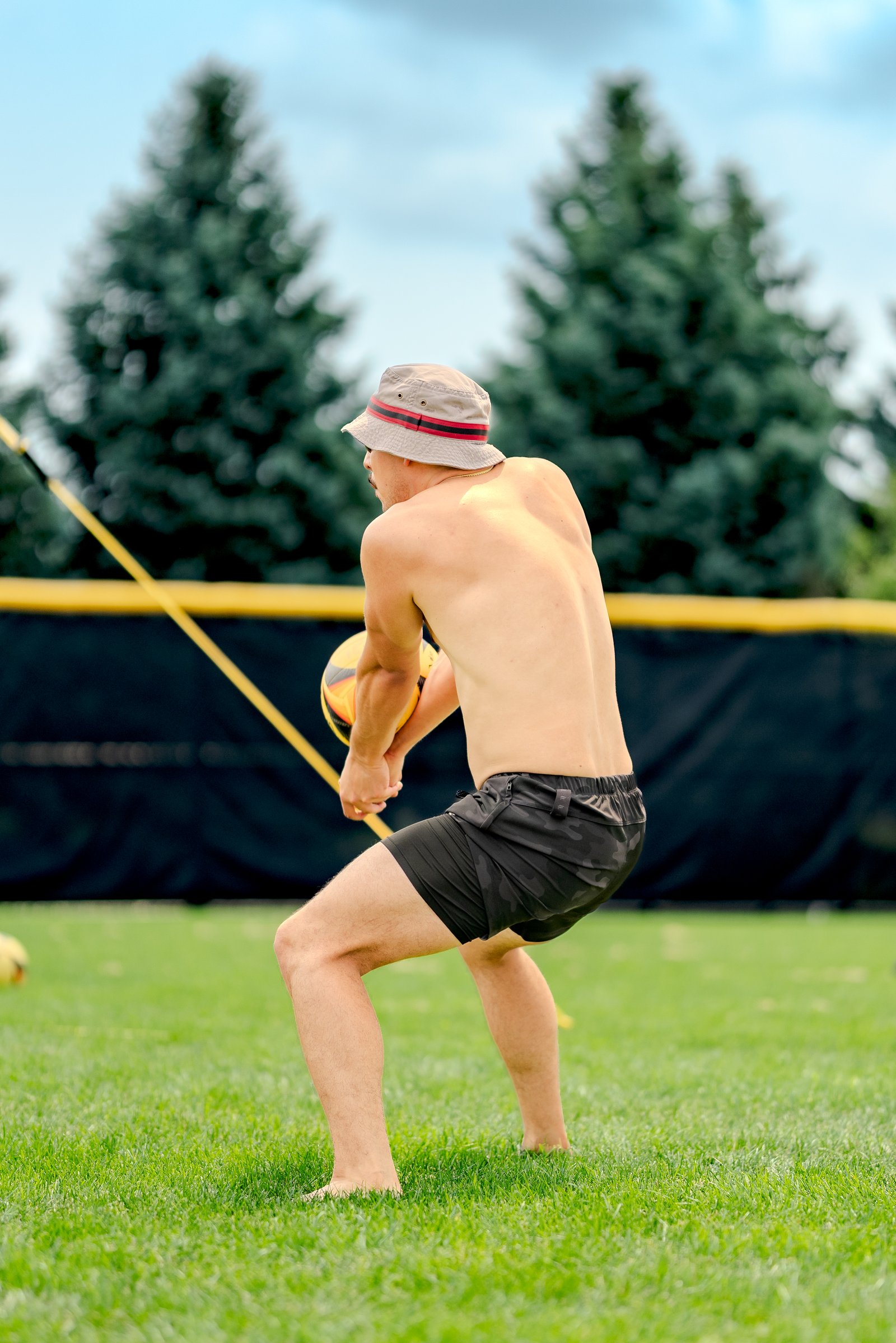 A player in black shorts and a bucket hat prepares to dig a volleyball on a grassy court with trees and a net in the background.