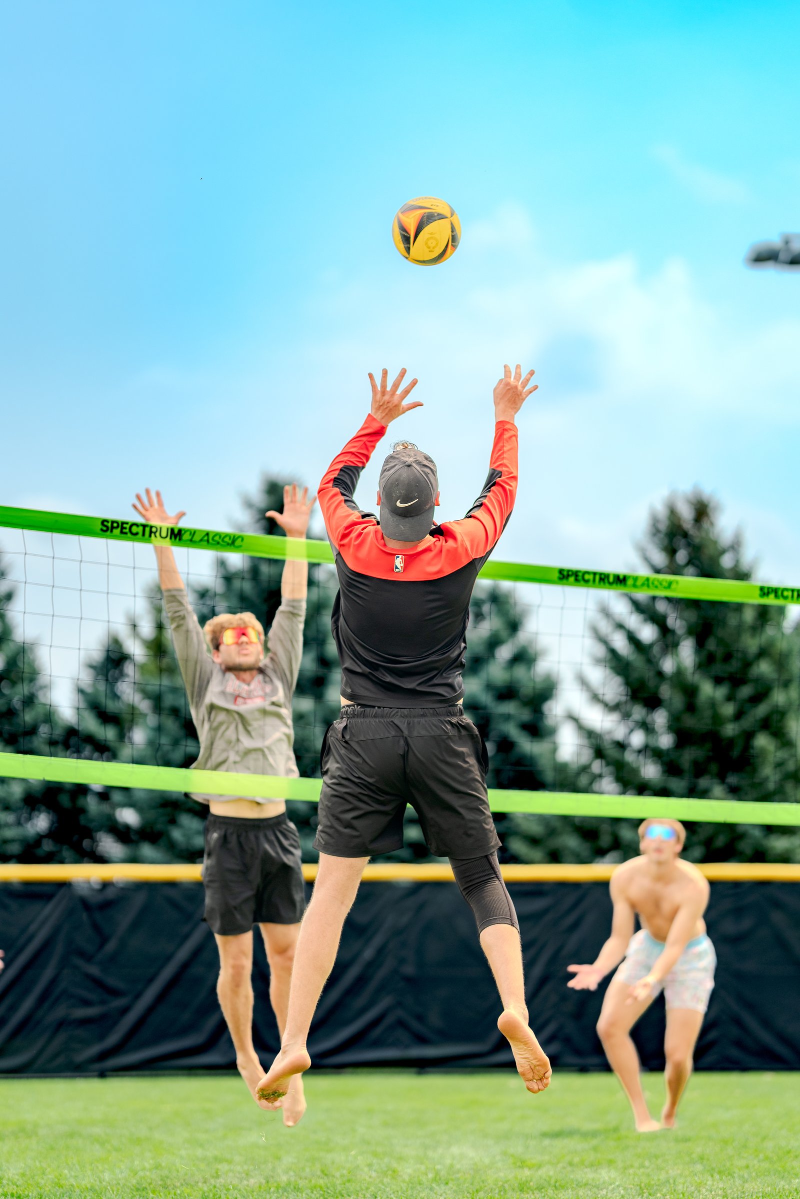 Players in black and gray tops jump near a volleyball net, with a player in patterned shorts in the background.