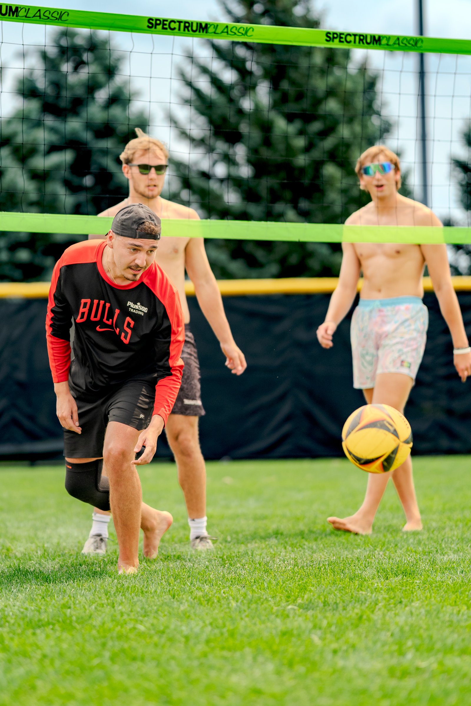 A player in a black and red top with 'BULLS' on it prepares to play volleyball, with two other players visible on the grass court.