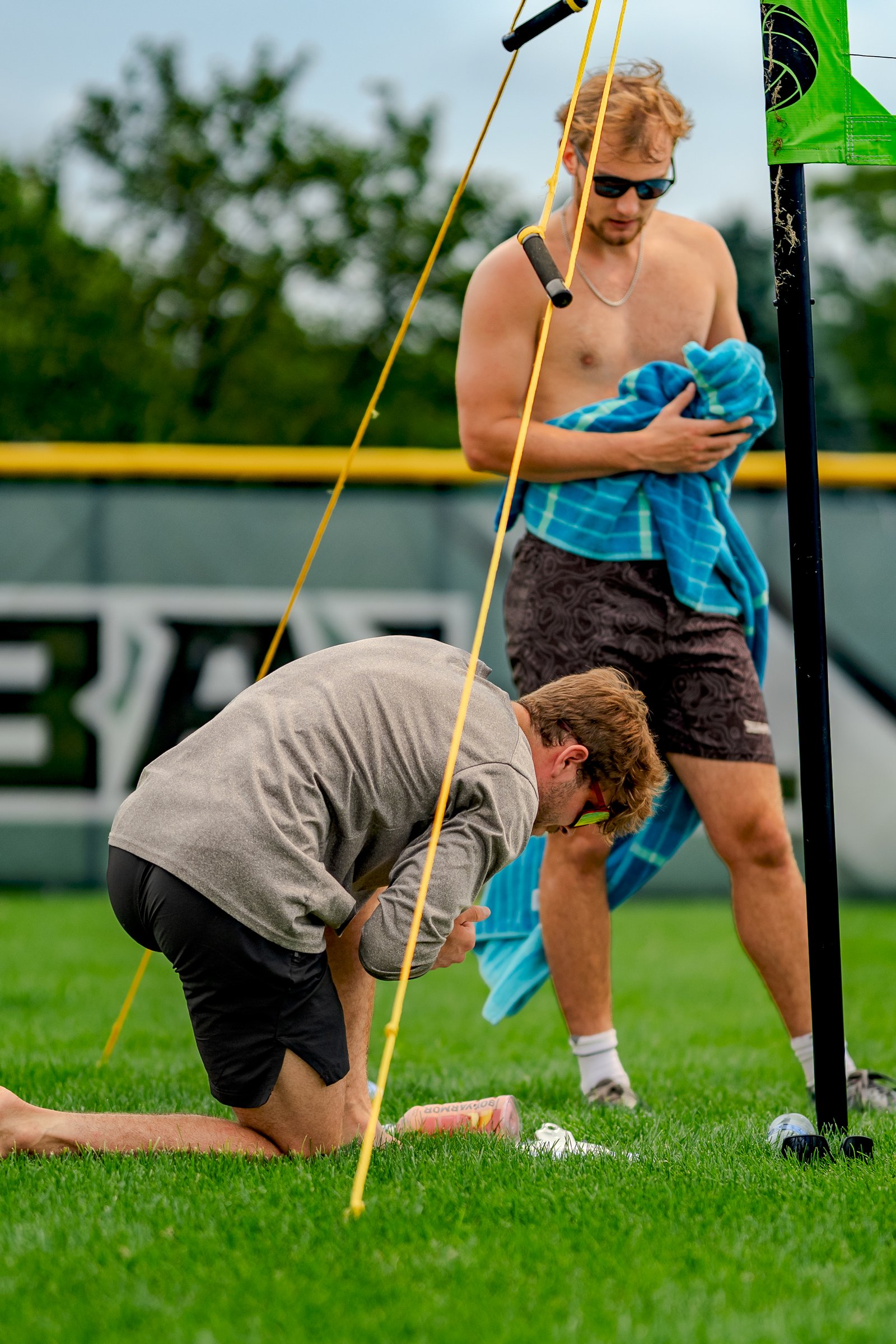 A player in a gray shirt and black shorts kneels on the grass, while another player in brown shorts holds a blue towel.