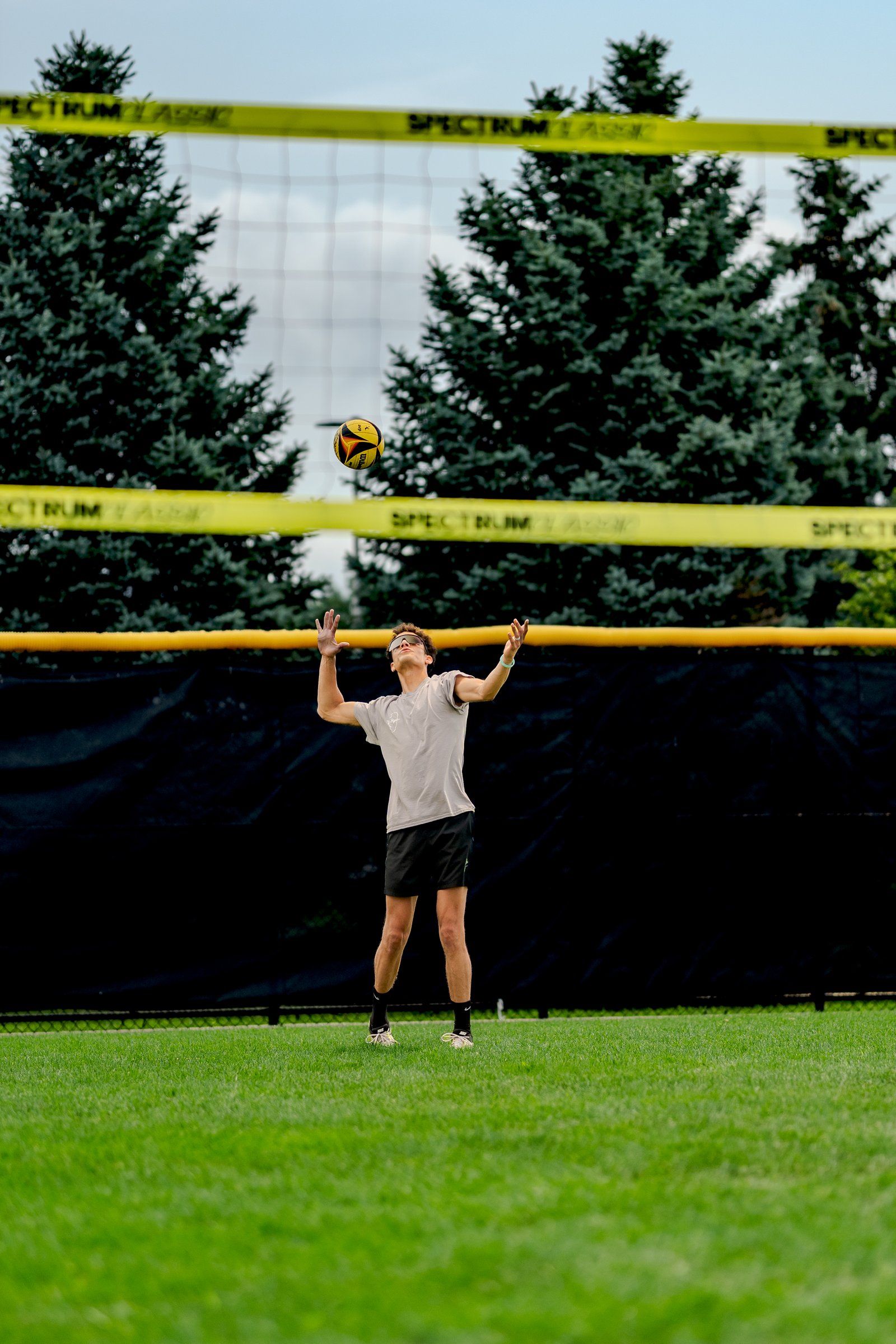A player in a gray top and black shorts sets a volleyball near the net with trees and a black fence in the background.