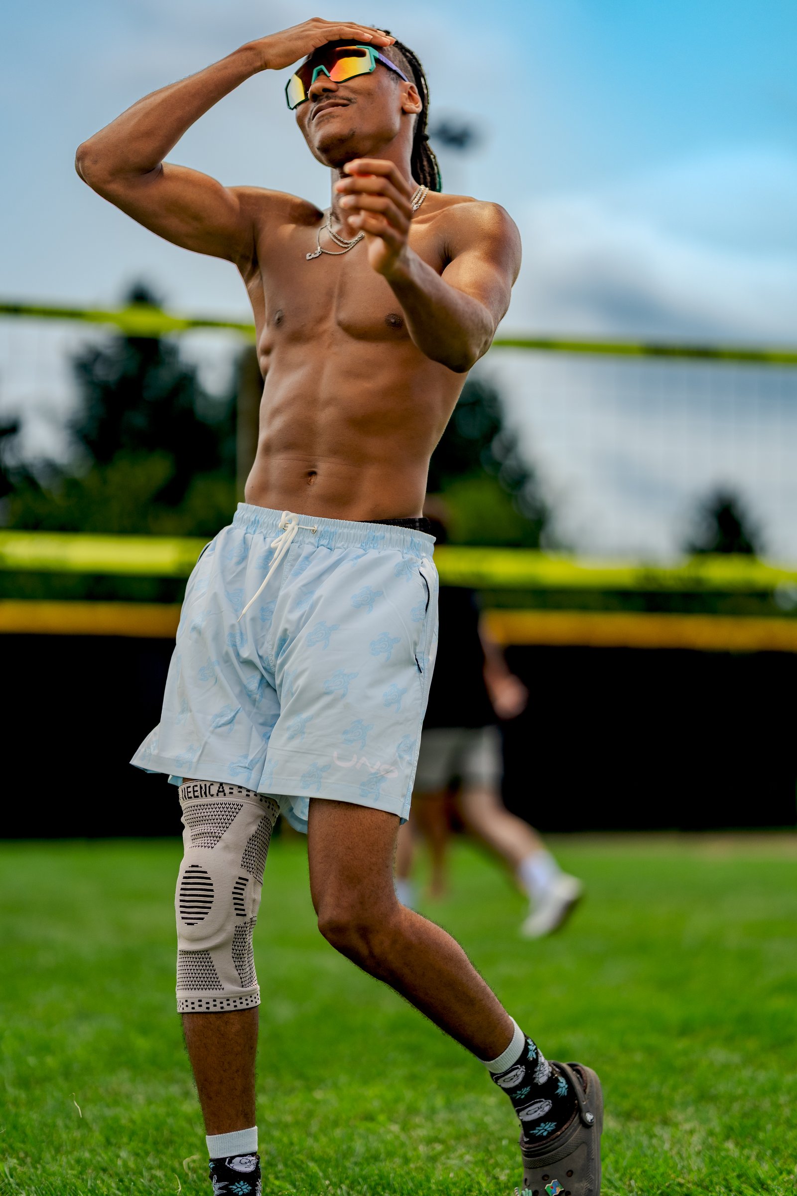 A player in light blue shorts with a turtle pattern shields his eyes from the sun on a grassy court with a volleyball net.