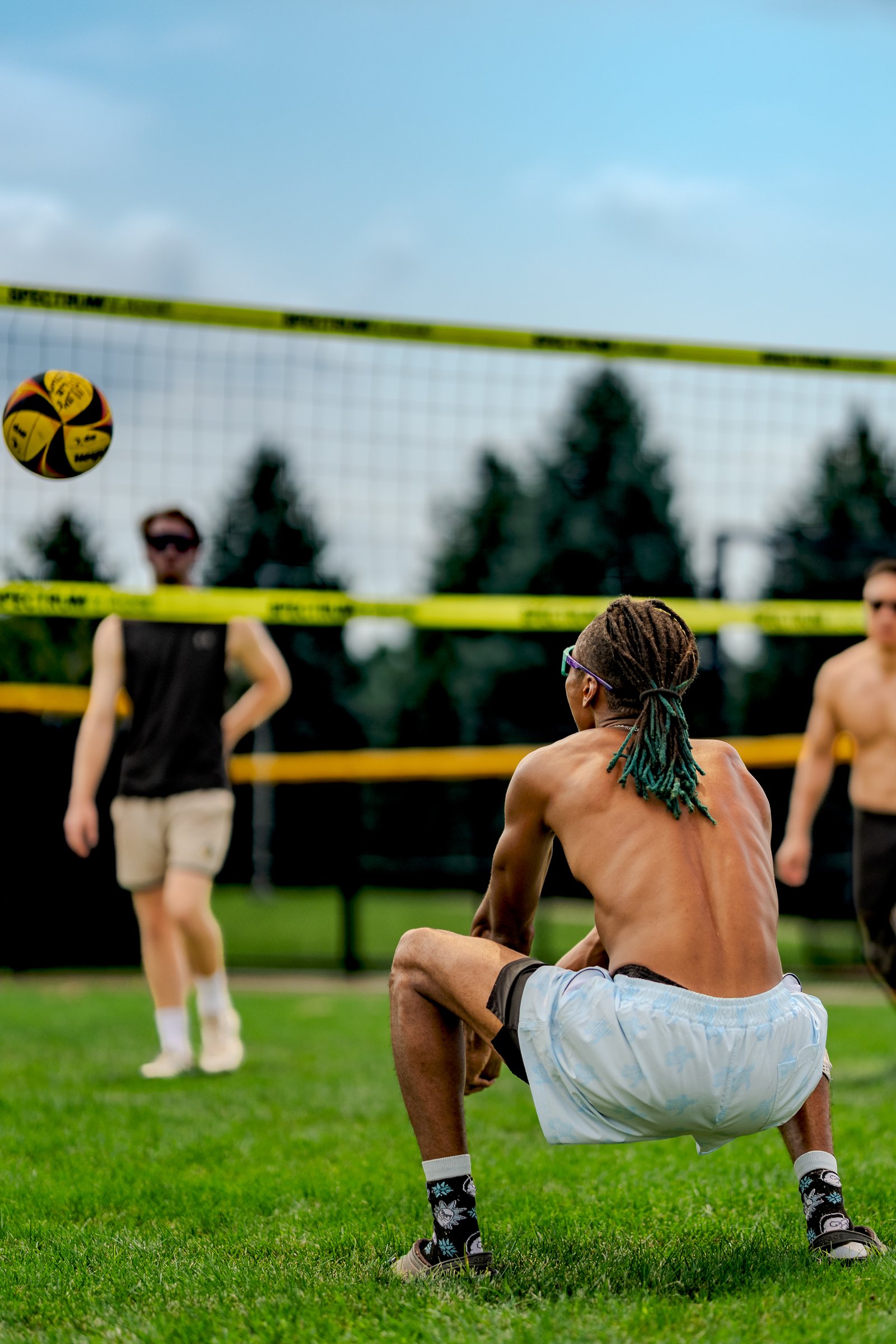 A player in light blue shorts crouches as a volleyball flies over the net, with players in black and shirtless in the background.