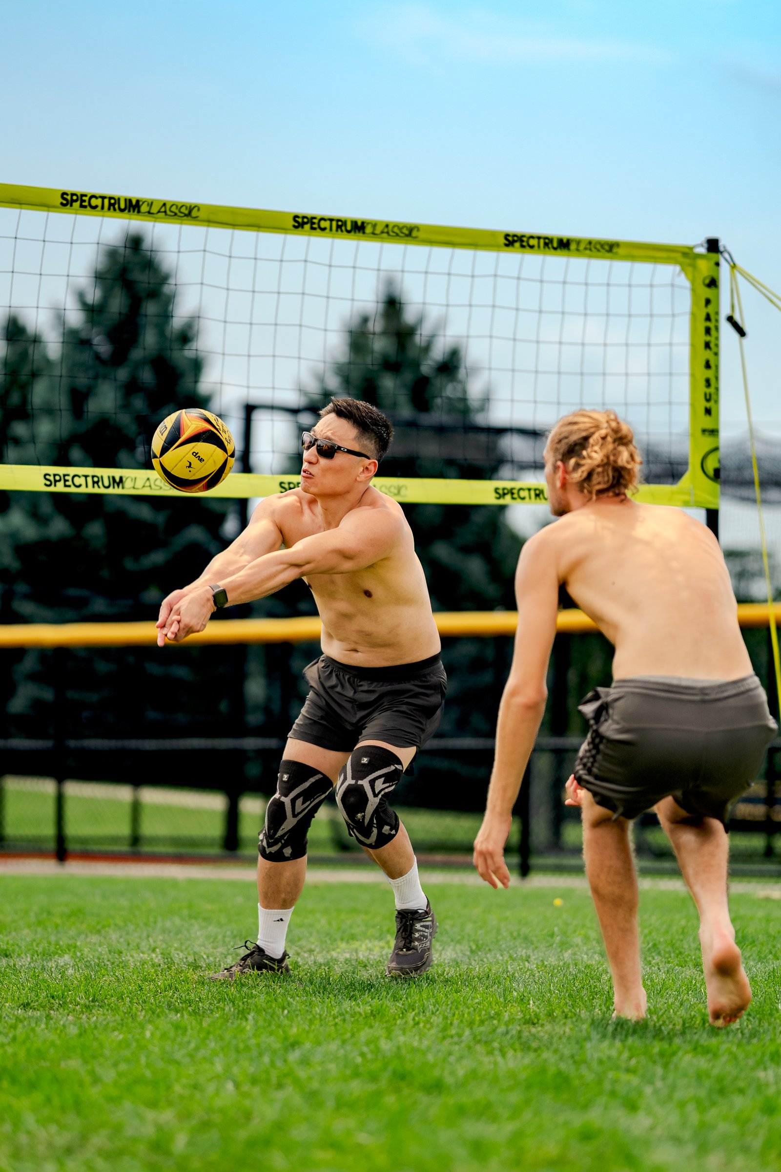 A player in black shorts passes a volleyball near a player in gray shorts on a grassy court with a net.