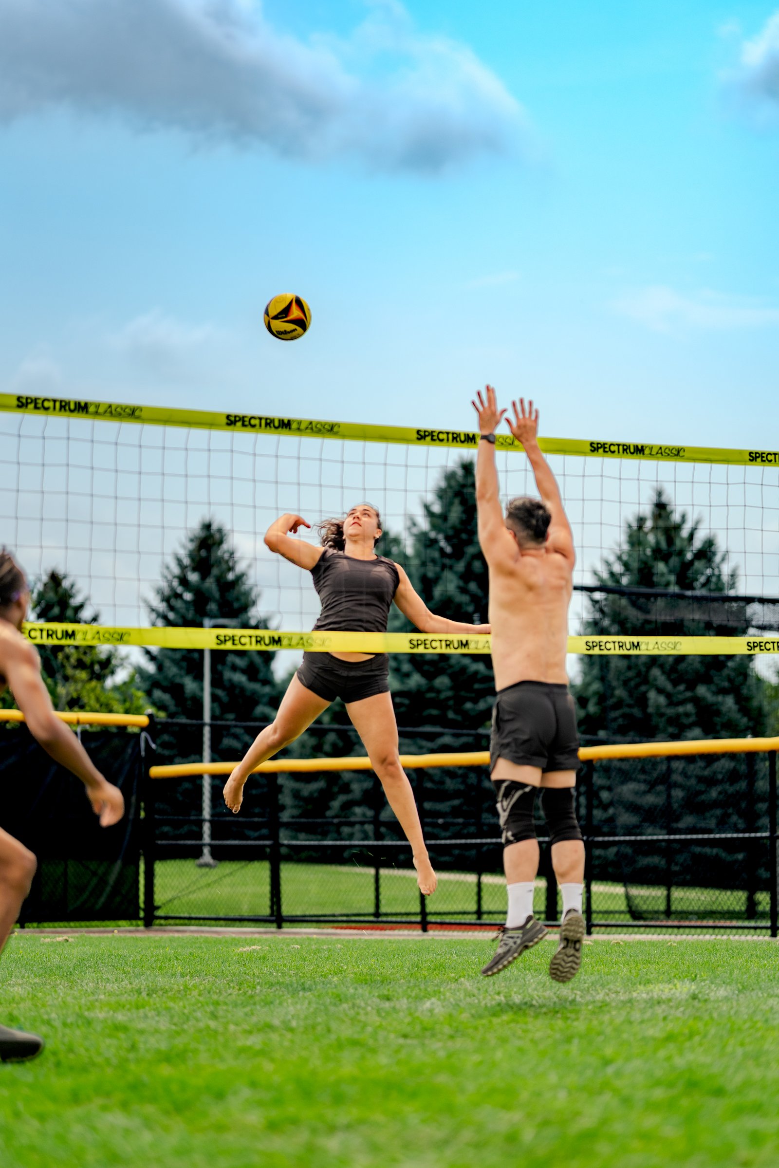 A player in a black top jumps to hit a volleyball over the net, with a player in black shorts reaching up to block.