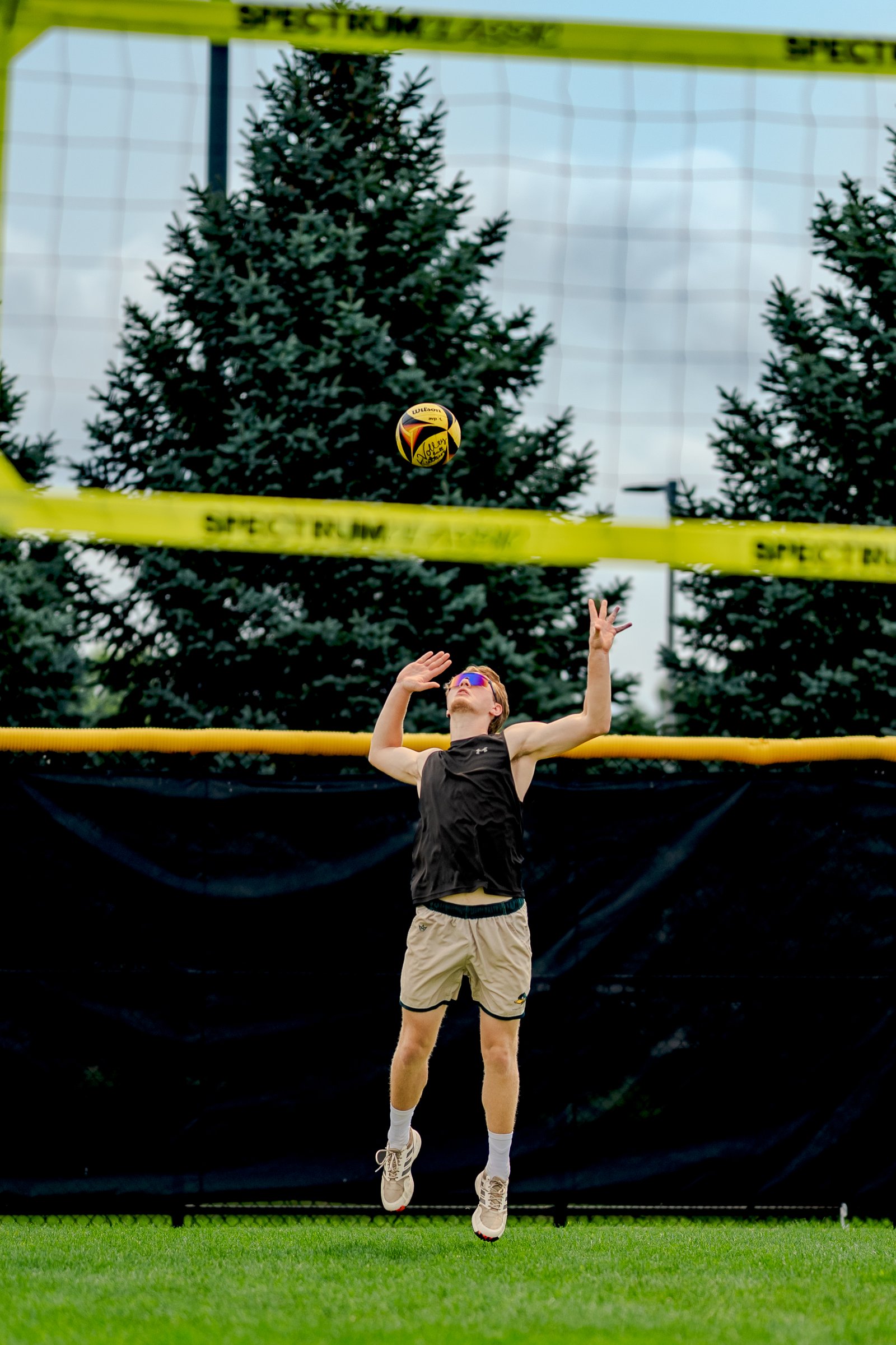 A player in a black top and tan shorts jumps to set a volleyball against a backdrop of trees and a net.