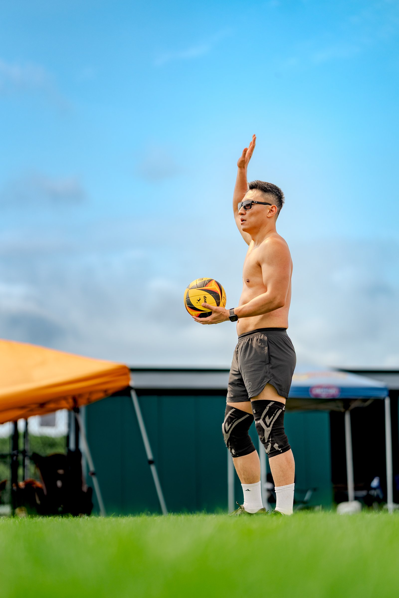 A player in black shorts and knee pads holds a volleyball, preparing to serve on a grassy court under a blue sky.