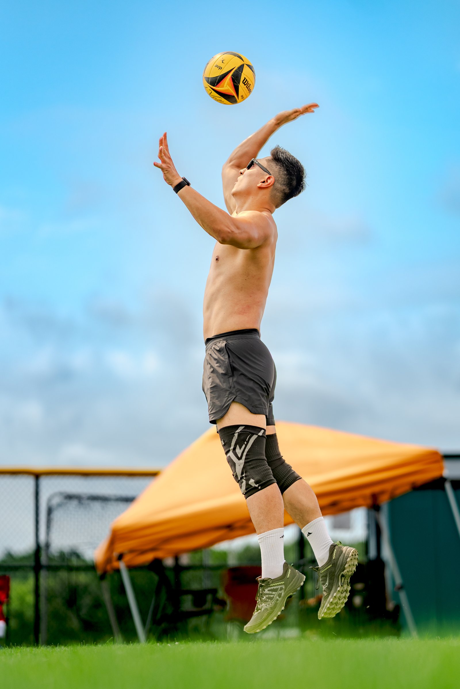 A player in black shorts and knee pads jumps to hit a volleyball against a blue sky with an orange tent in the background.