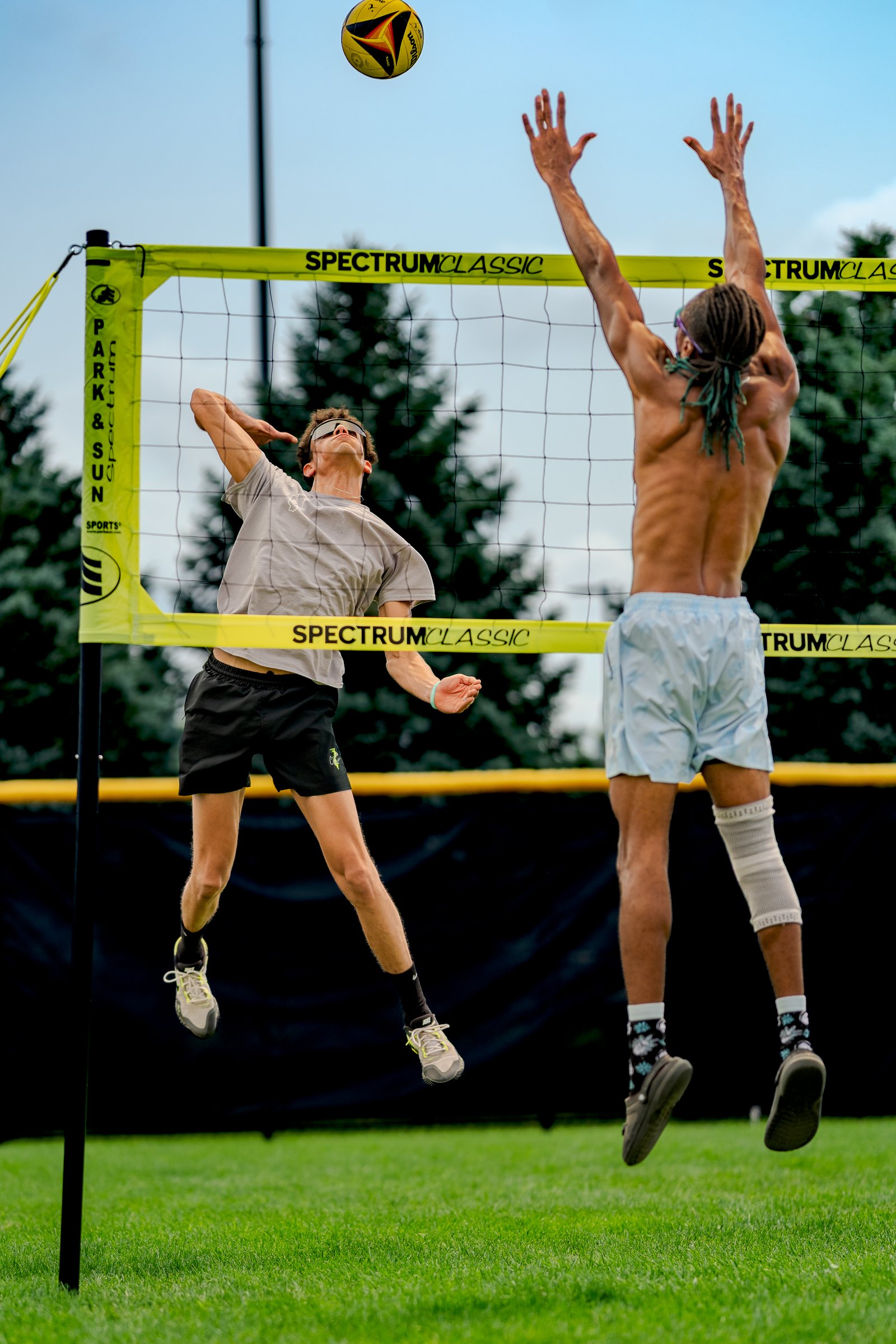 A player in a grey shirt and black shorts jumps to hit a volleyball, while a player in light blue shorts reaches to block.