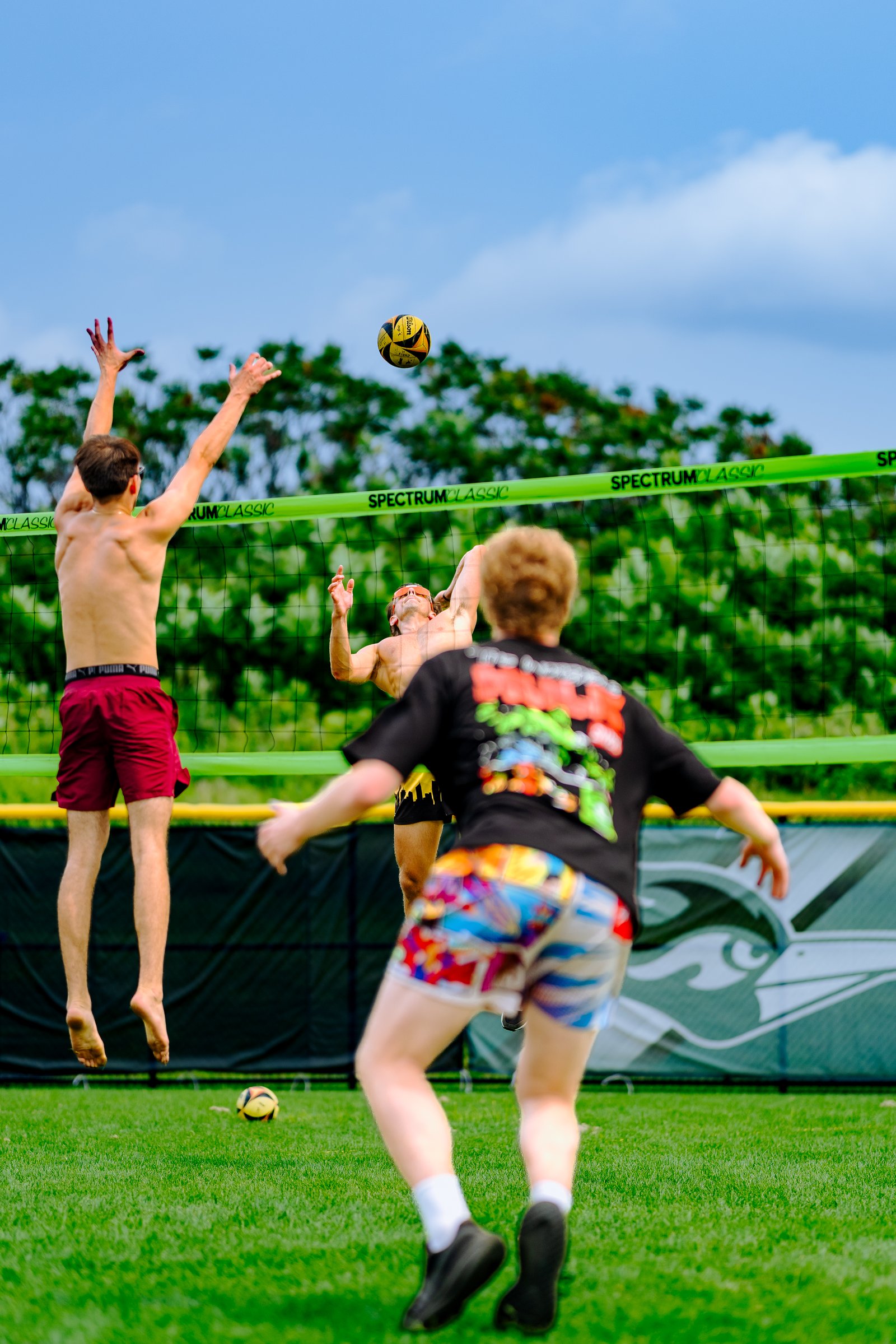 Players in red shorts and a black shirt jump near the net as a volleyball is in the air on a grassy court.
