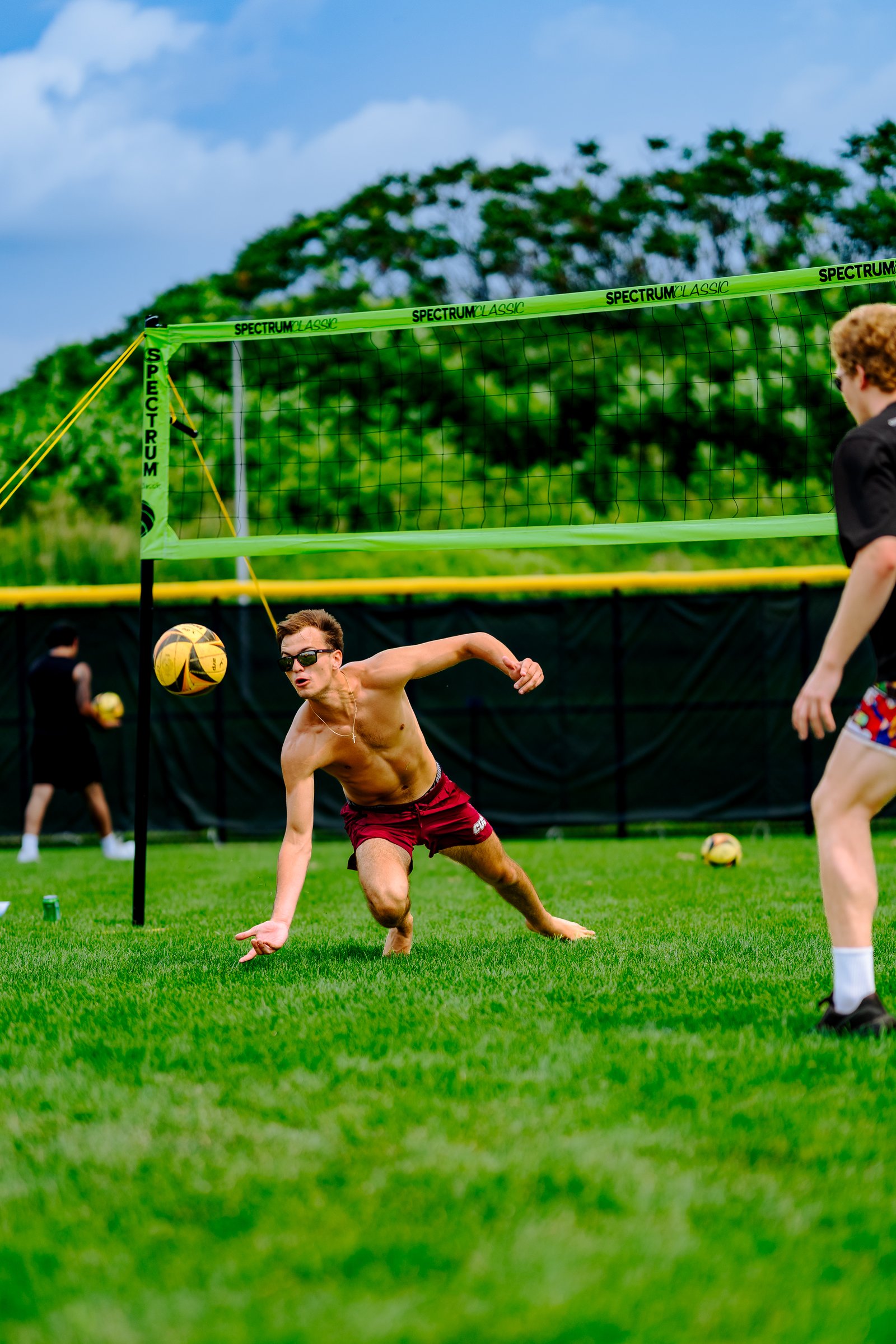 A player in maroon shorts dives to dig a volleyball on a grassy court with a green net and trees in the background.