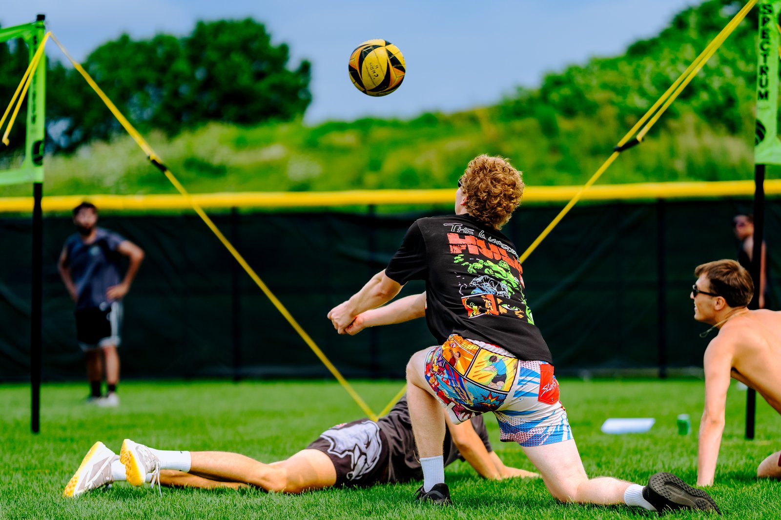 A player in a black shirt and colorful shorts attempts a dig as a yellow ball flies overhead, with other players on the grass court.