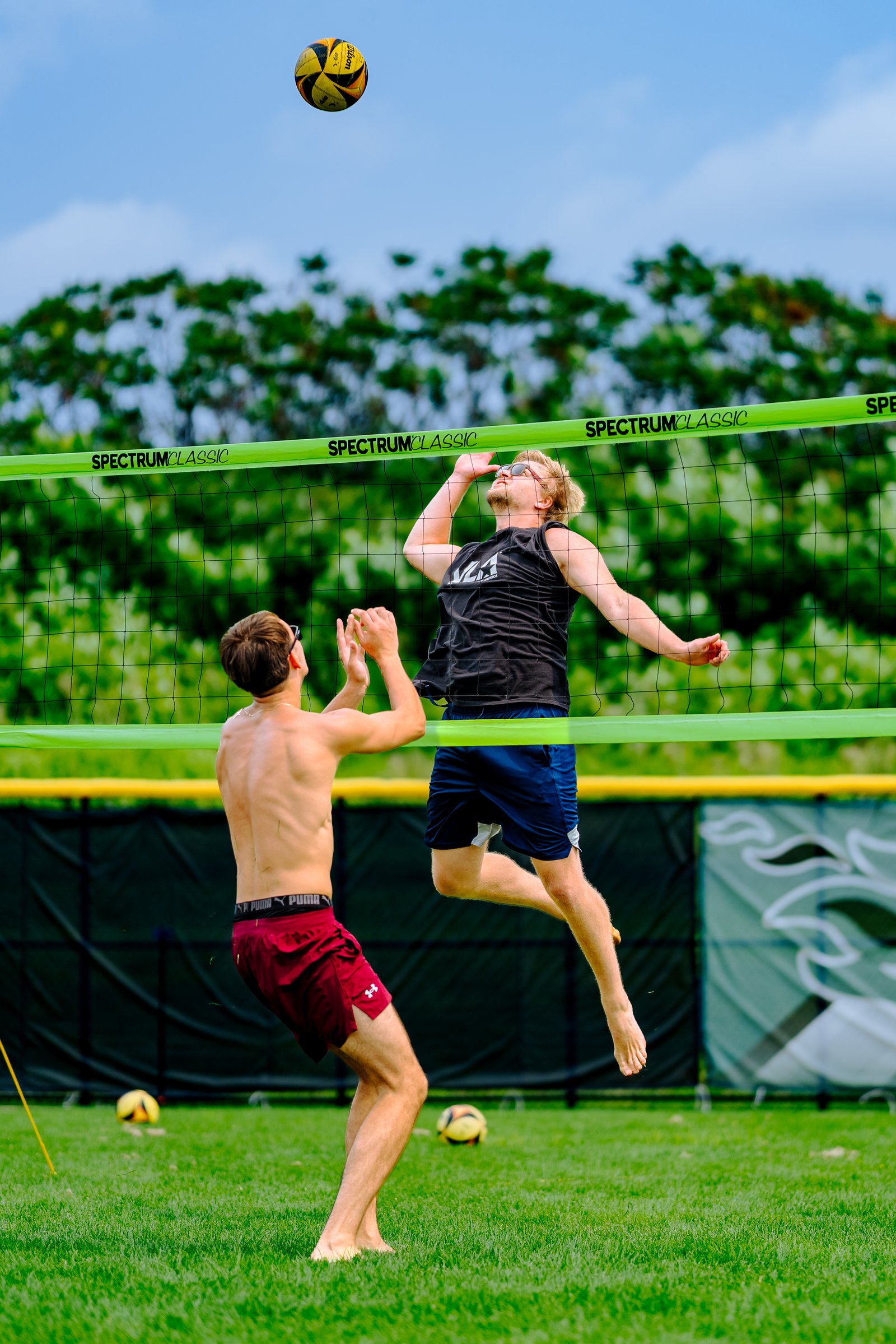 A player in a black top jumps to hit a volleyball over the net, while a player in maroon shorts prepares to receive.