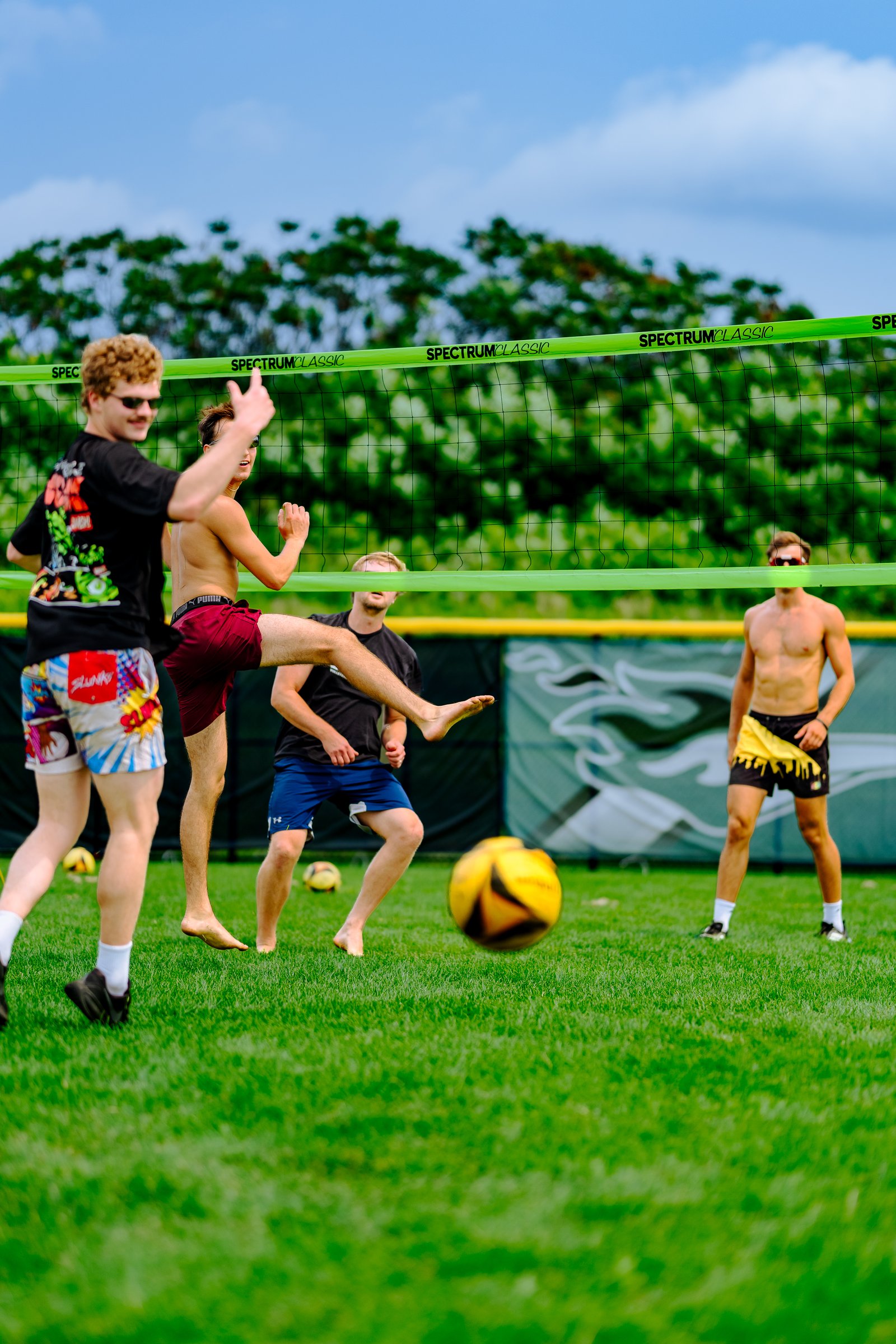 A player in maroon shorts kicks a yellow ball near the net, with other players in black tops and colorful shorts visible on the grass.