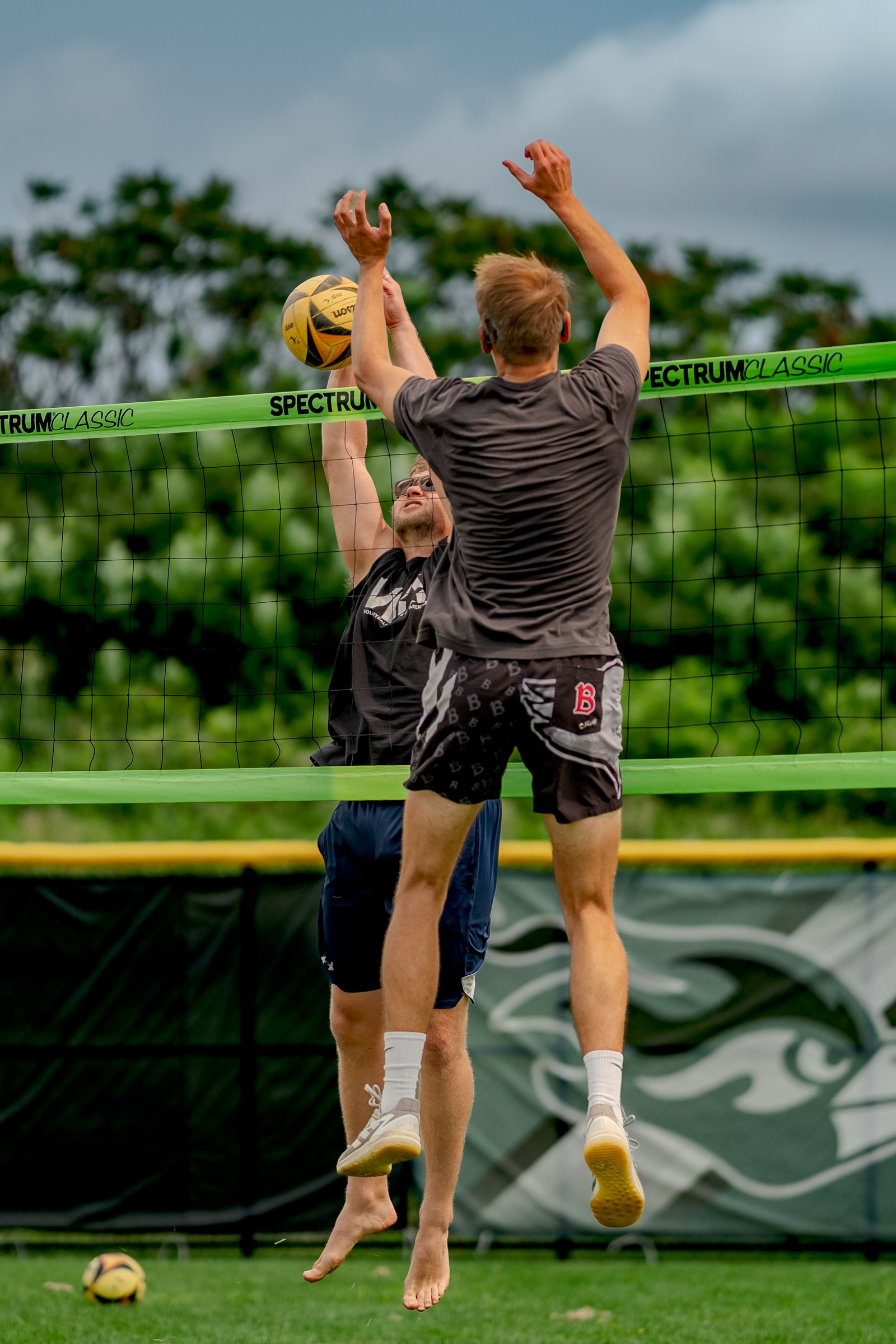 Two players jump at the net, one in a black top and navy shorts, the other in a grey top and patterned shorts, reaching for the ball.
