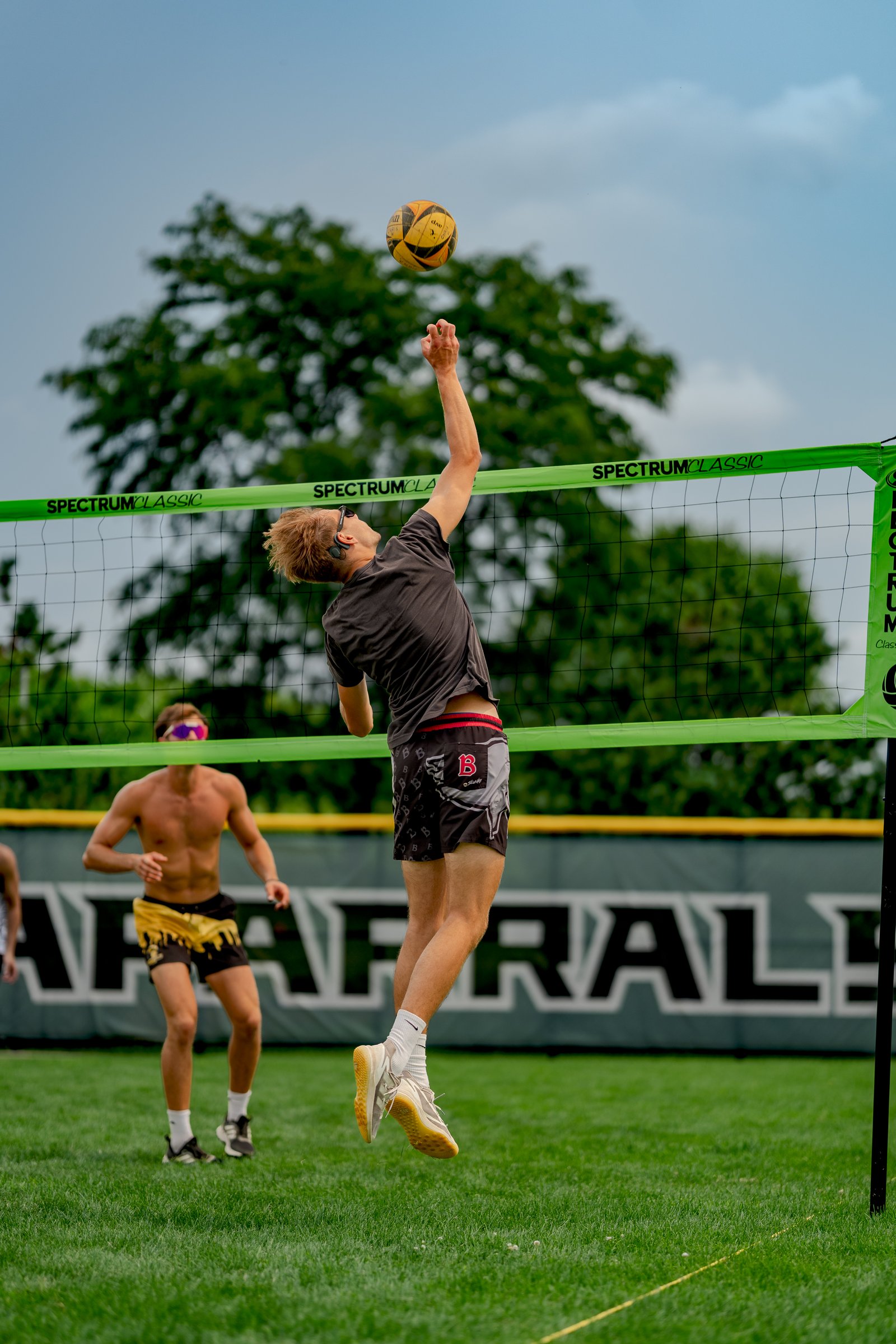 A player in a dark shirt and patterned shorts jumps to hit a volleyball over the net, with another player in yellow shorts watching.