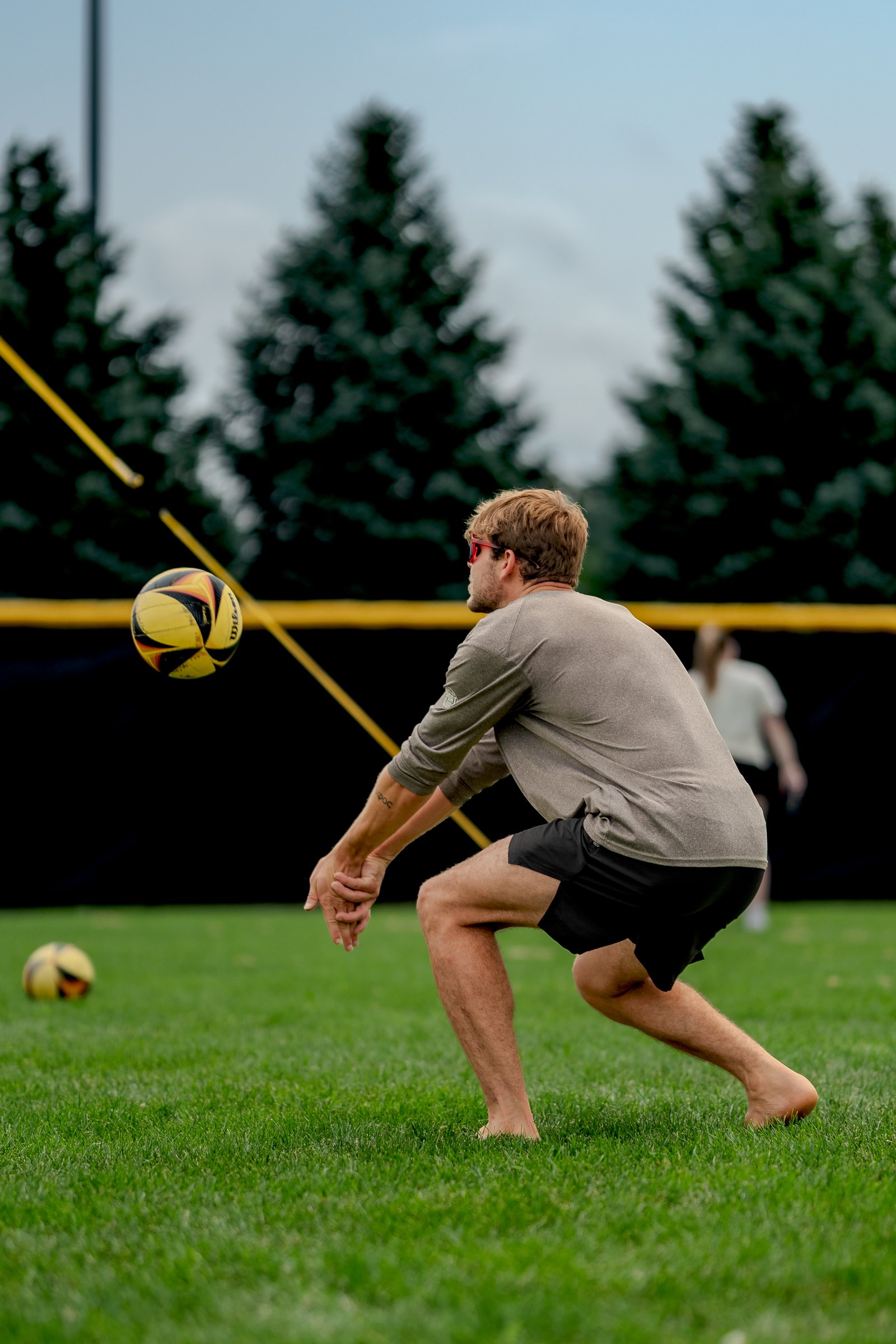 A player in a gray long-sleeve shirt and black shorts prepares to dig a volleyball on a grassy court with trees in the background.