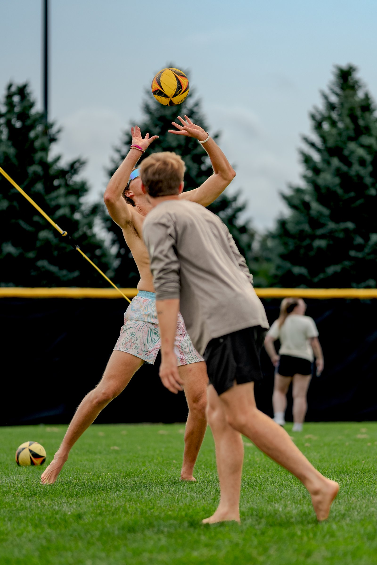 A player in patterned shorts reaches for a volleyball as another player in a gray shirt moves forward on the grass court.