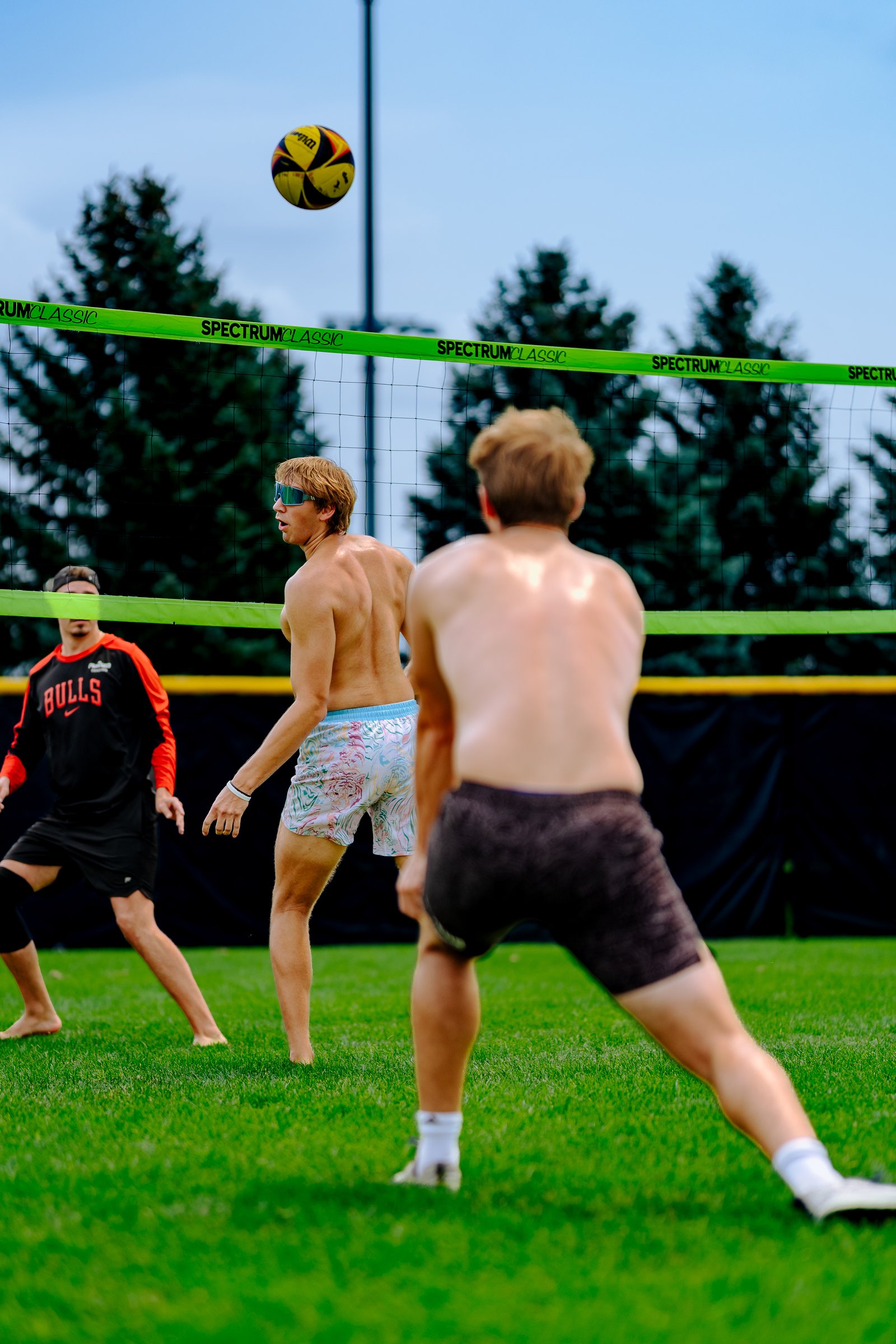 A player in patterned shorts prepares to hit a volleyball over the net, with a player in a black and red Bulls jersey nearby.