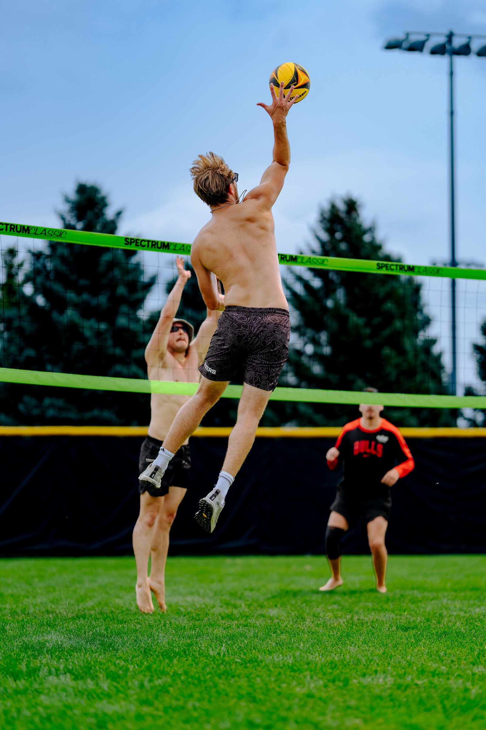A player in black shorts jumps to hit a volleyball over the net, with two other players visible on the grass court.