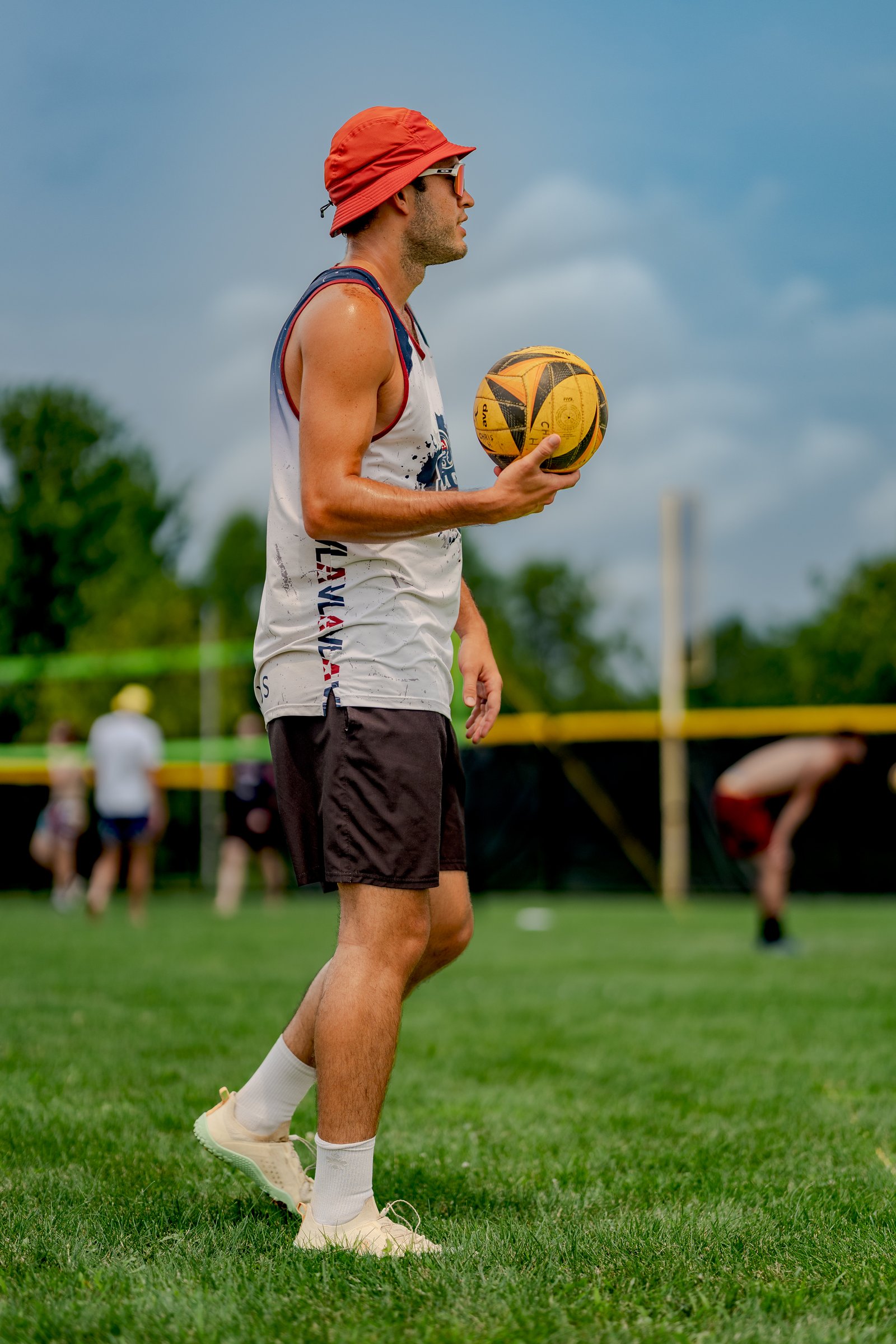 A player in a white and blue jersey holds a volleyball on a grassy court with other players in the background.