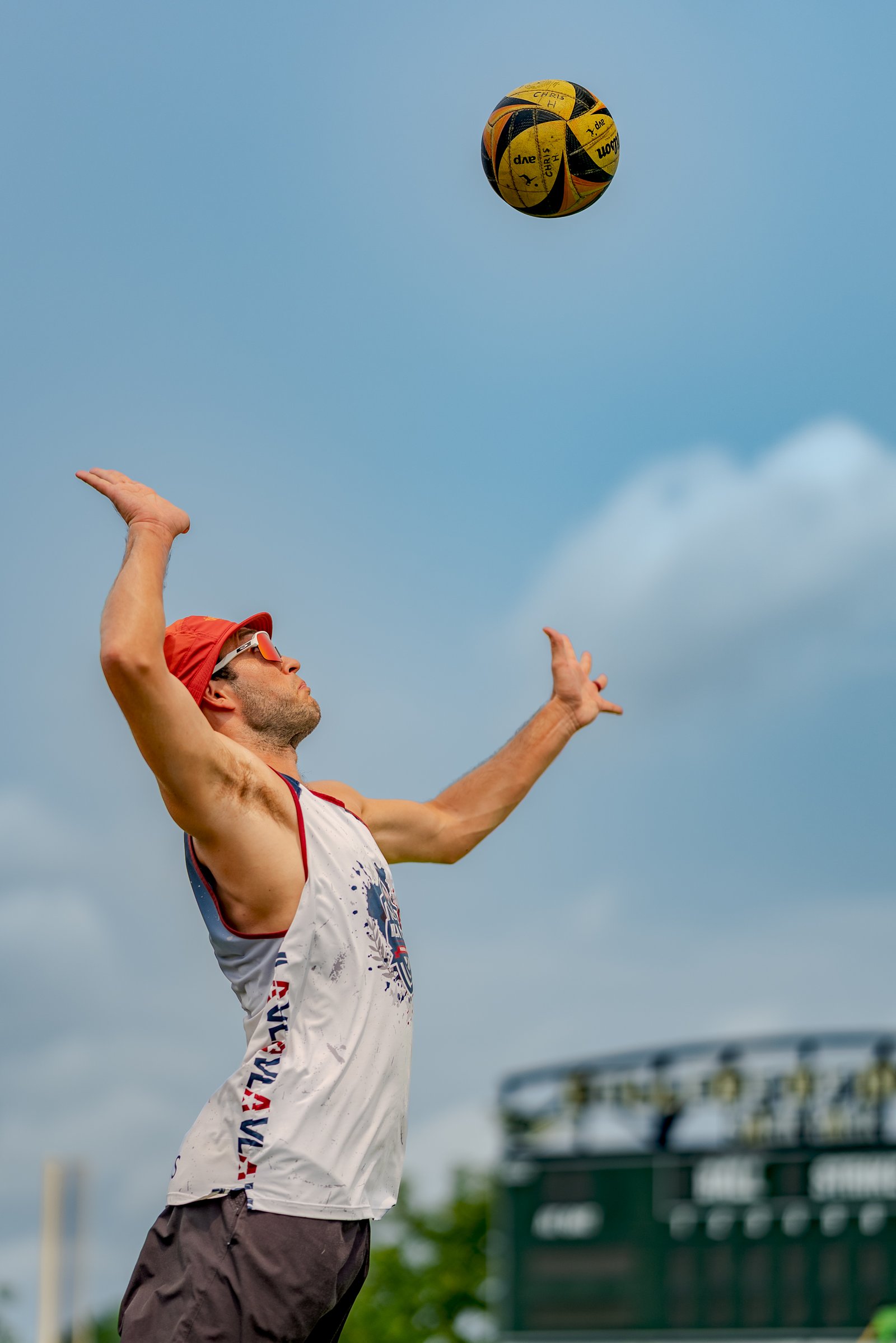 A player in a white jersey with red and blue trim serves a volleyball against a blue sky.