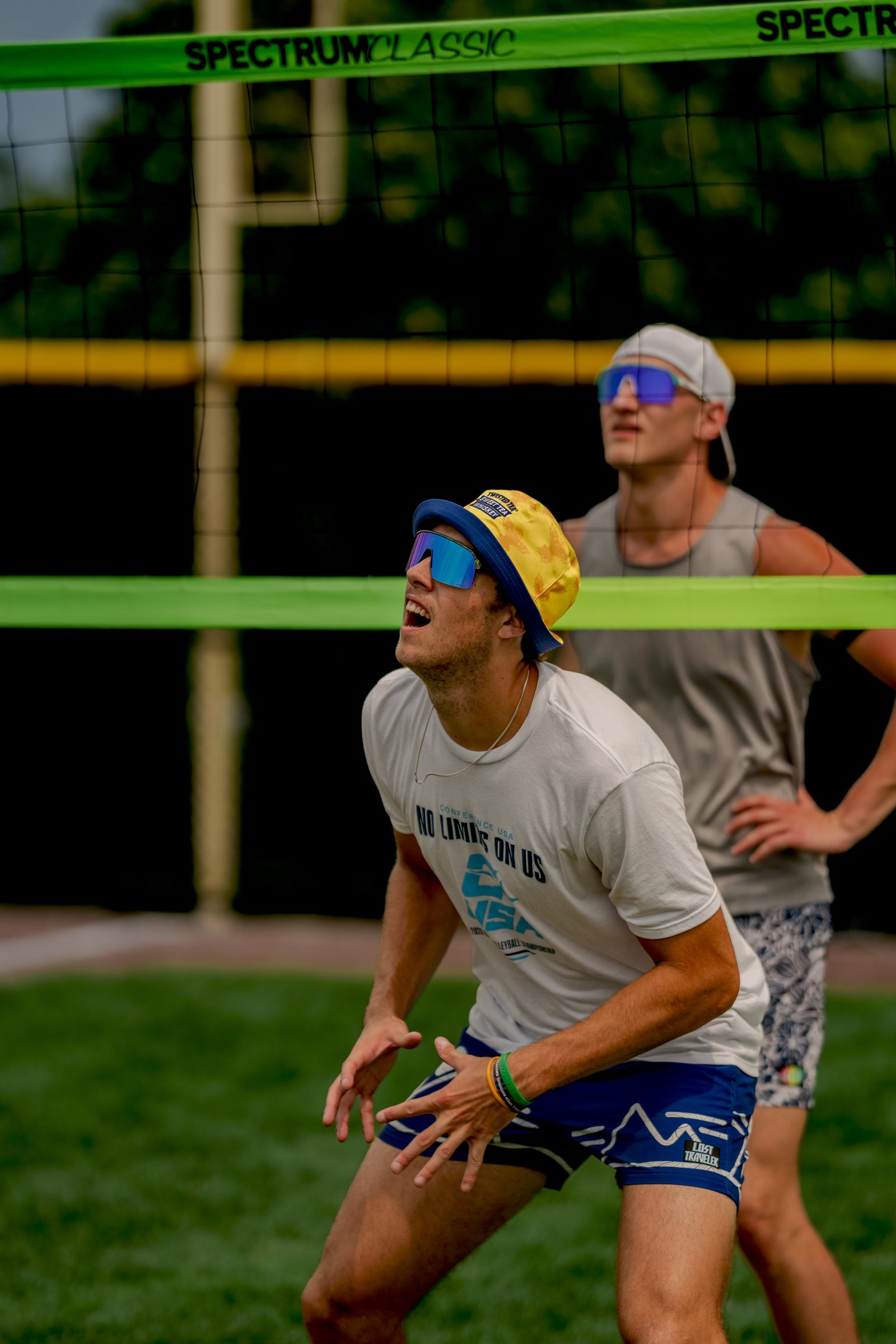 A player in a white shirt and blue shorts prepares to play volleyball, with another player in a gray tank top behind him.