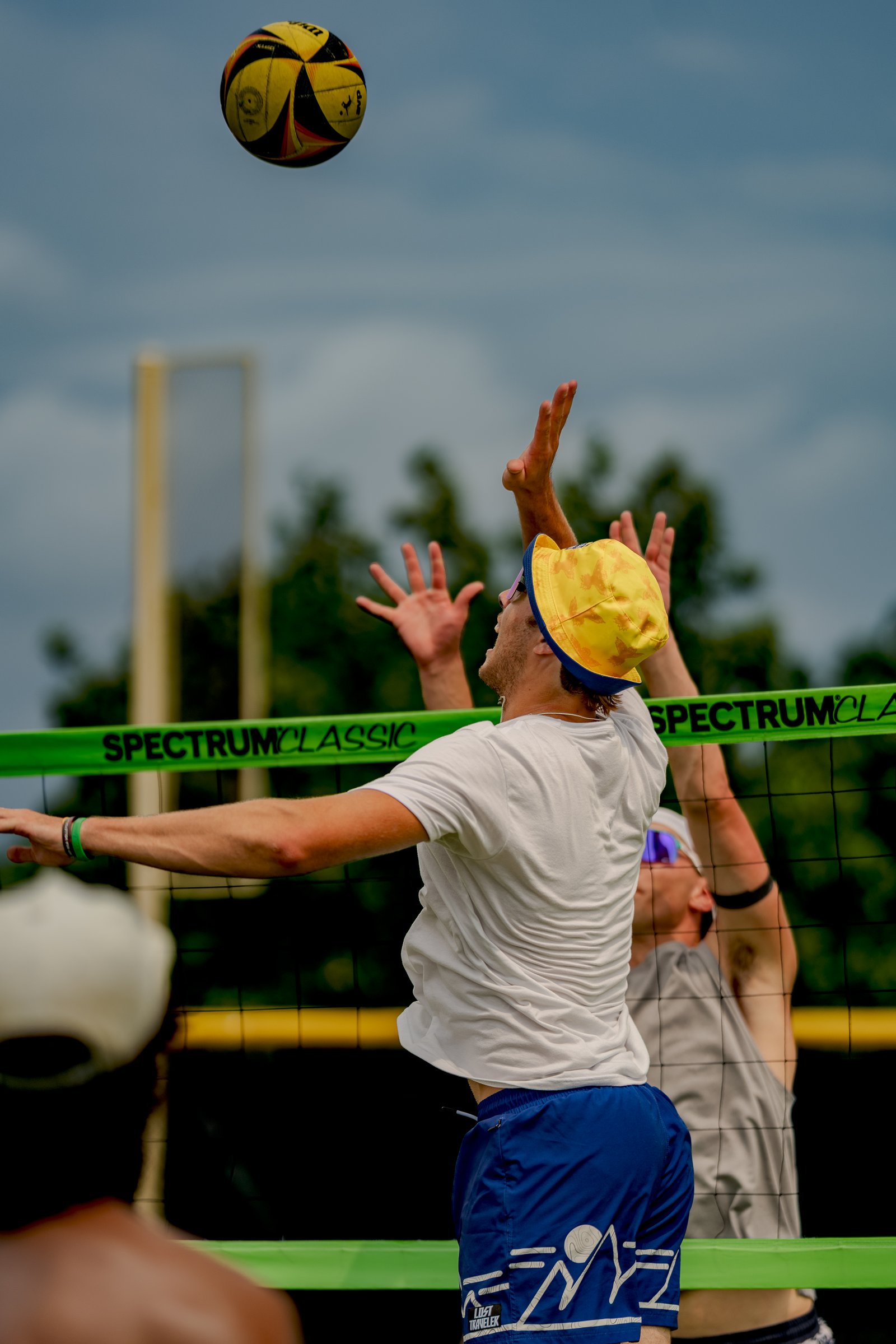 A player in blue shorts and a white shirt jumps to hit a volleyball over the net, with another player in a gray shirt reaching up.
