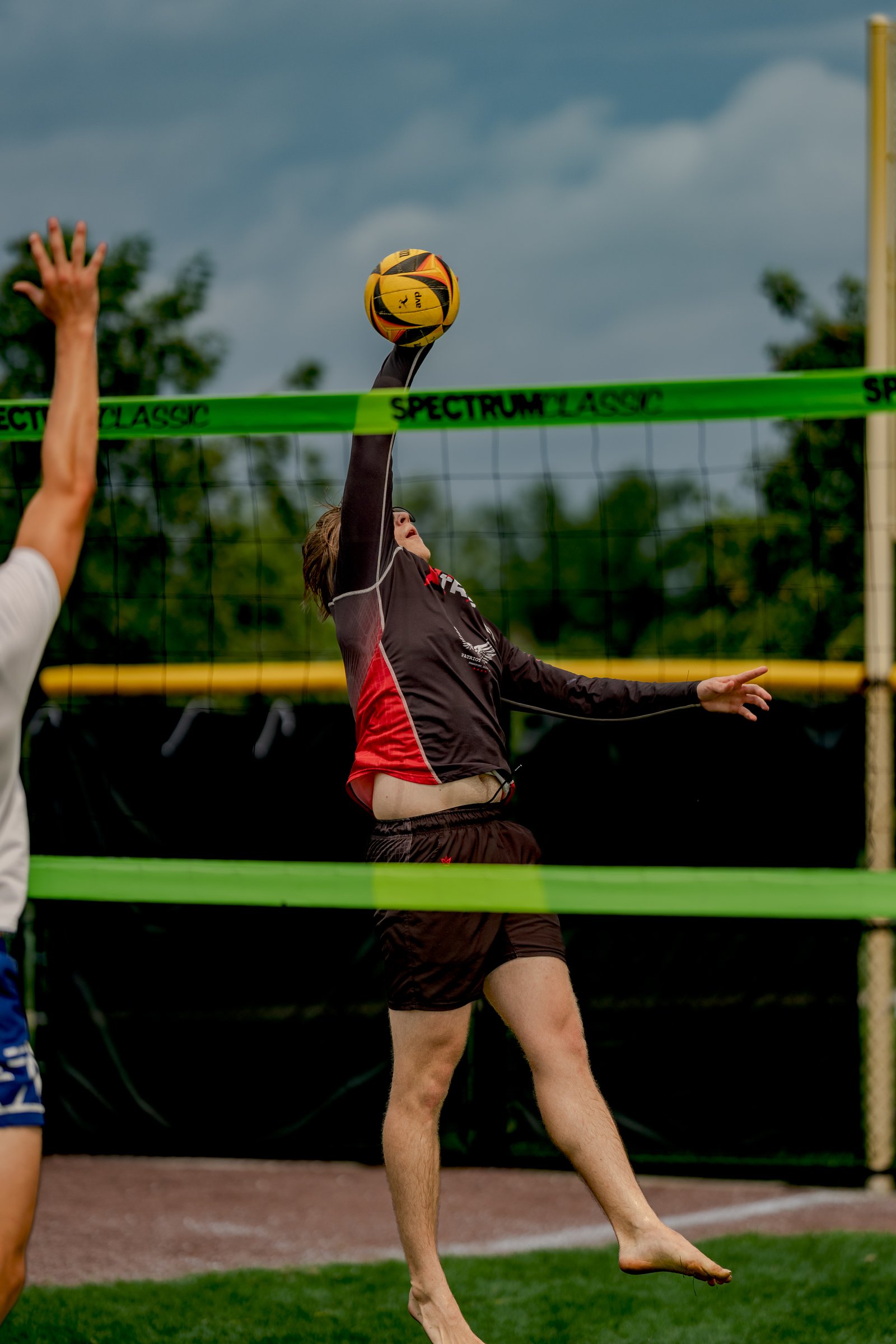 A player in a black and red top spikes a volleyball over the net, with another player in a white top reaching up.