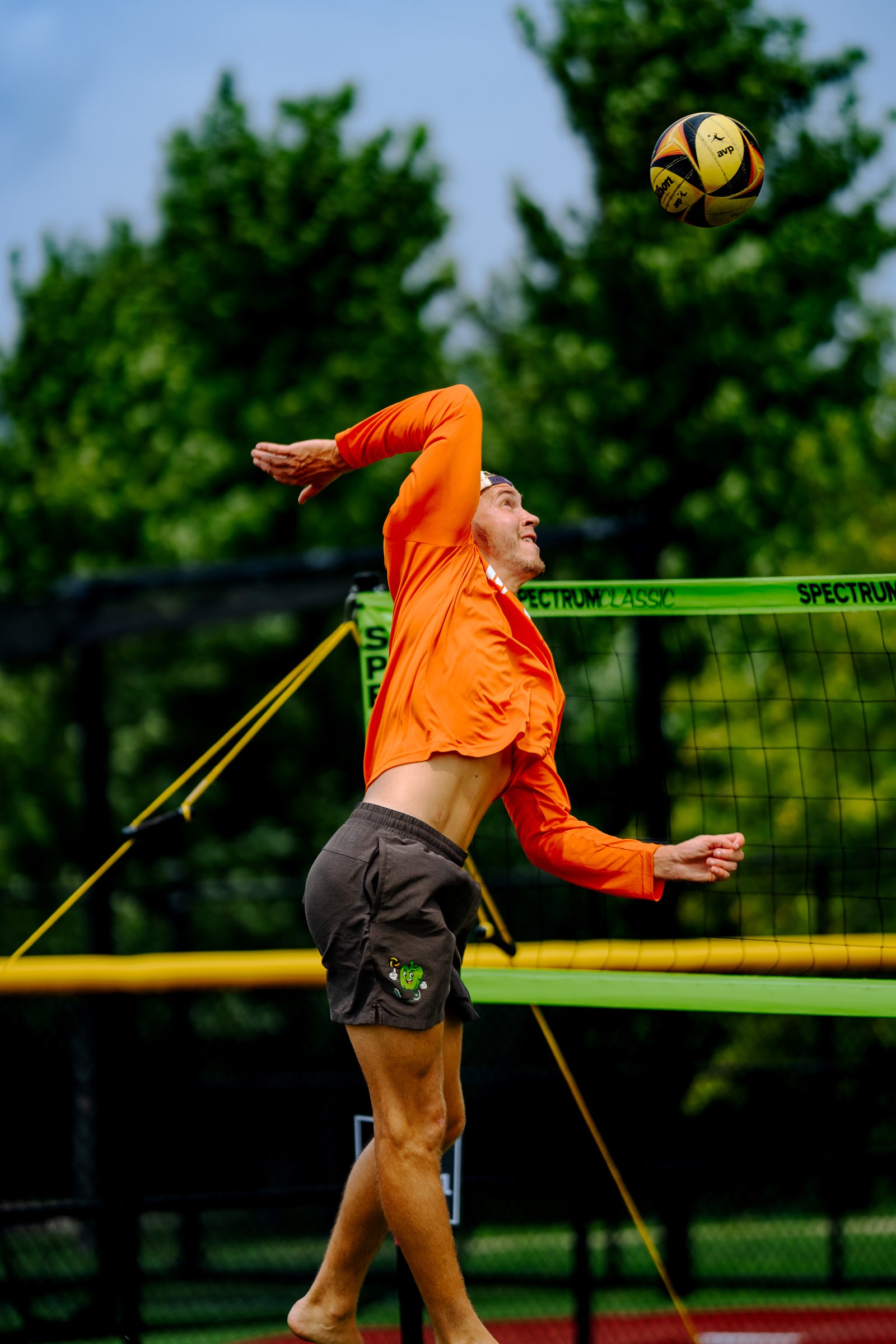 A player in an orange shirt and dark shorts jumps to hit a volleyball over the net.