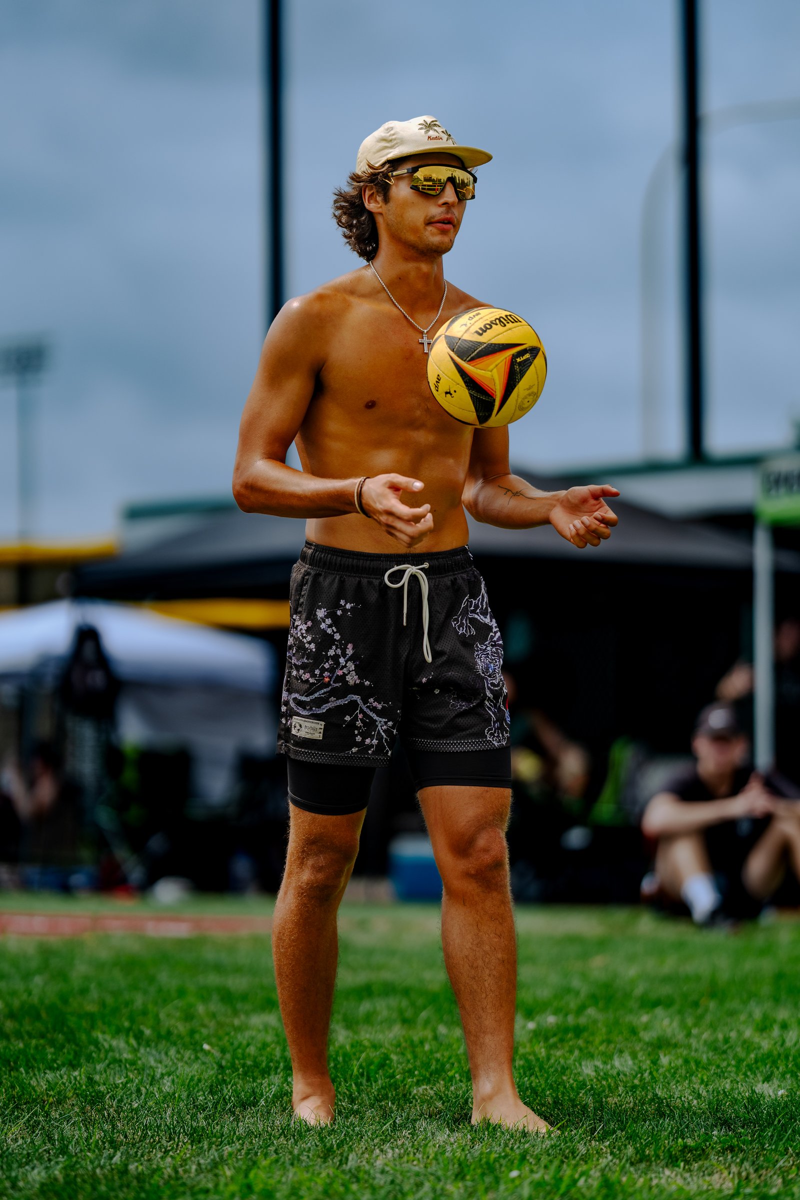 A player in black shorts with a floral pattern tosses a yellow volleyball in the air on a grassy court.