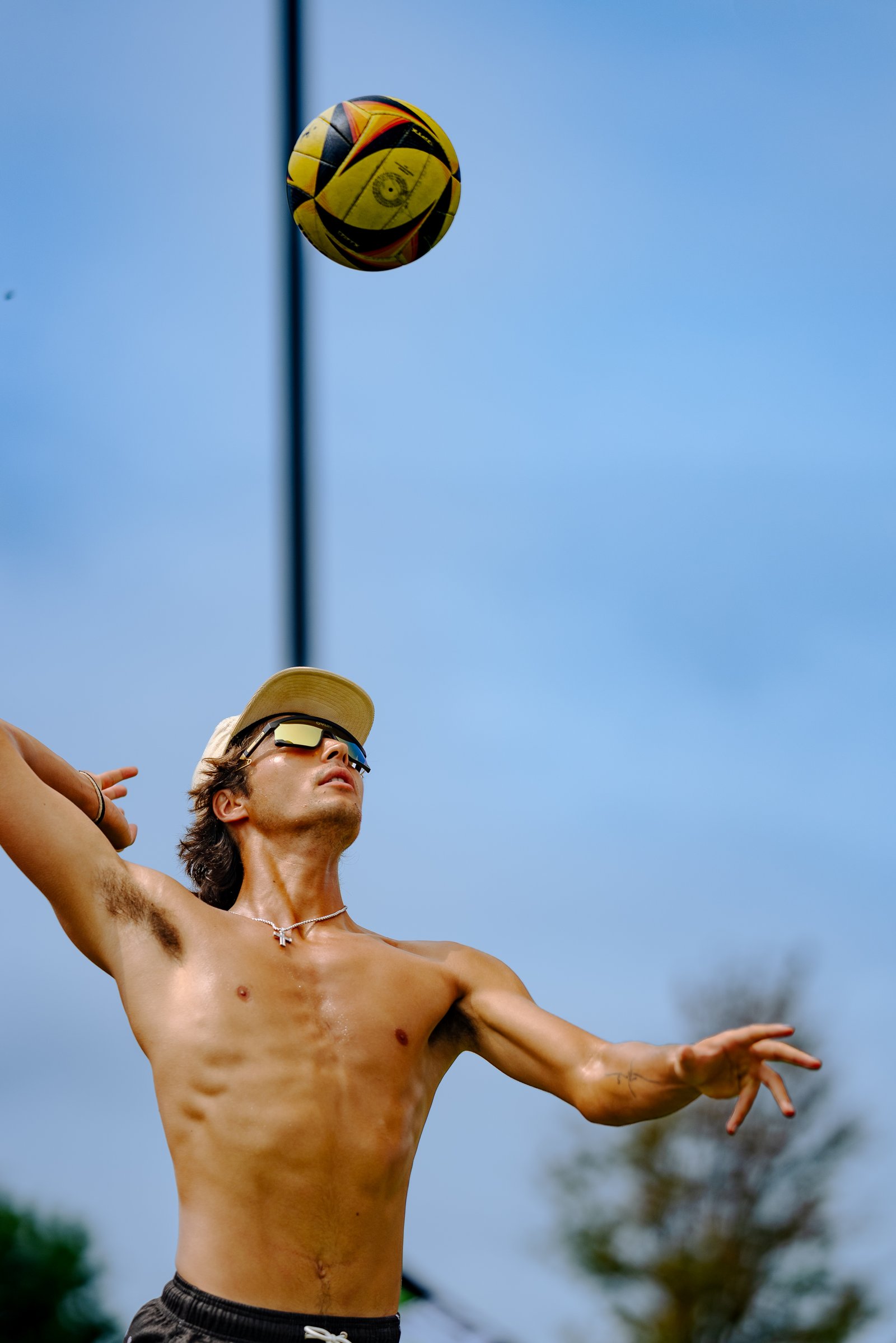 A player in black shorts and a tan cap with yellow sunglasses prepares to hit a yellow and black volleyball against a blue sky.