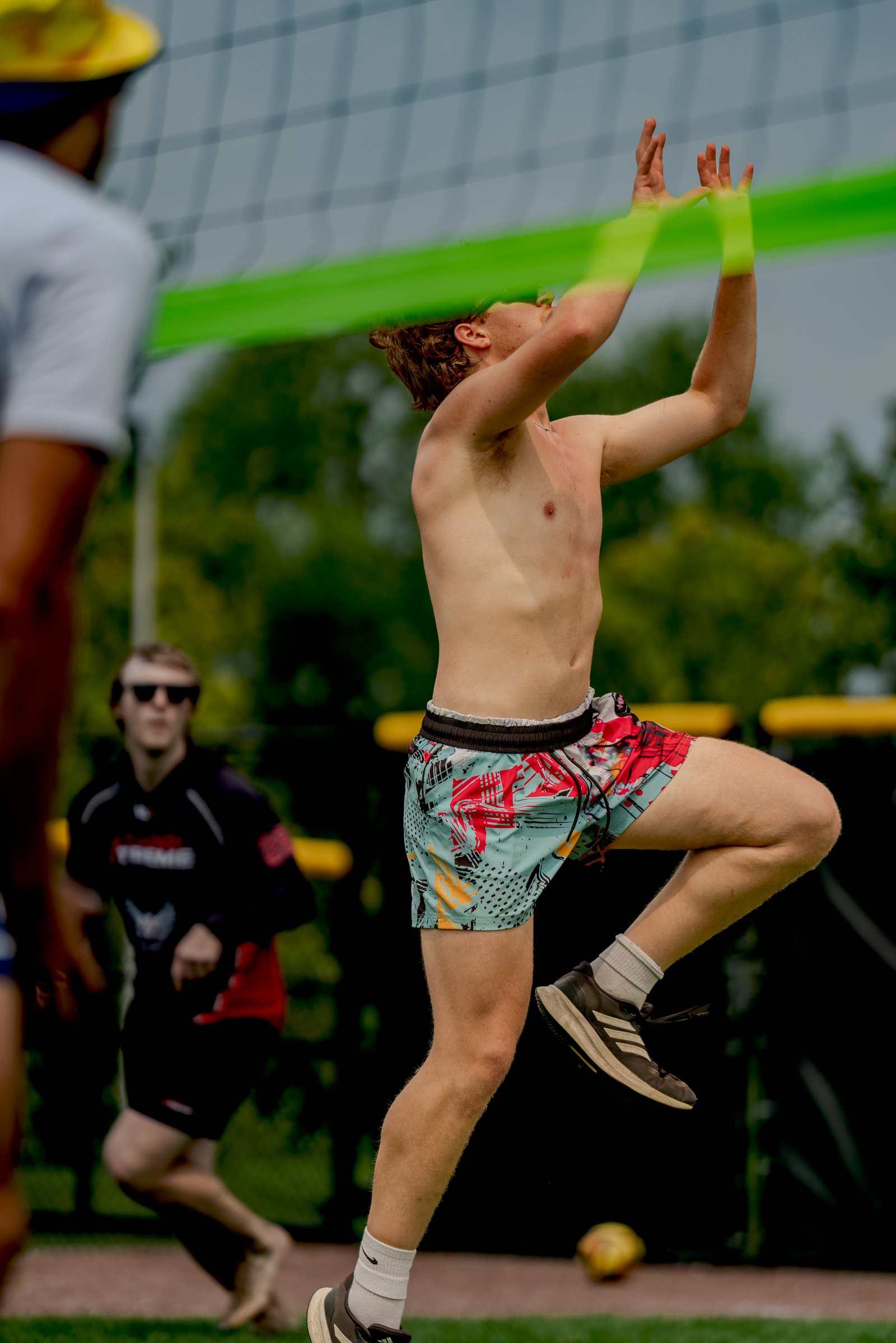 A player in colorful shorts jumps to set the ball near the net, with other players in black and red visible in the background.