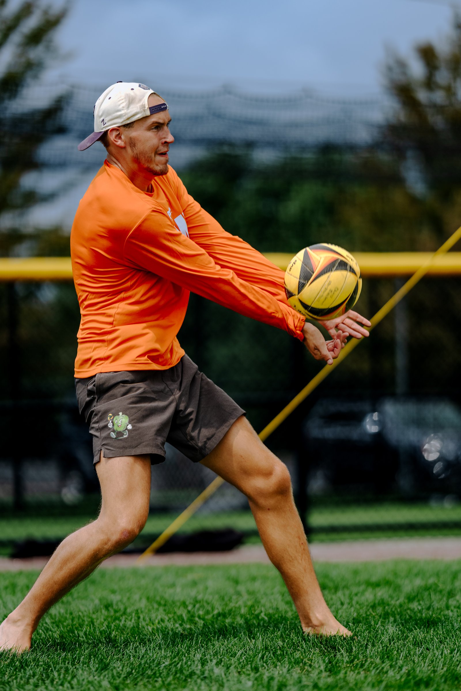 A player in an orange shirt and gray shorts passes a yellow volleyball on a grassy court.