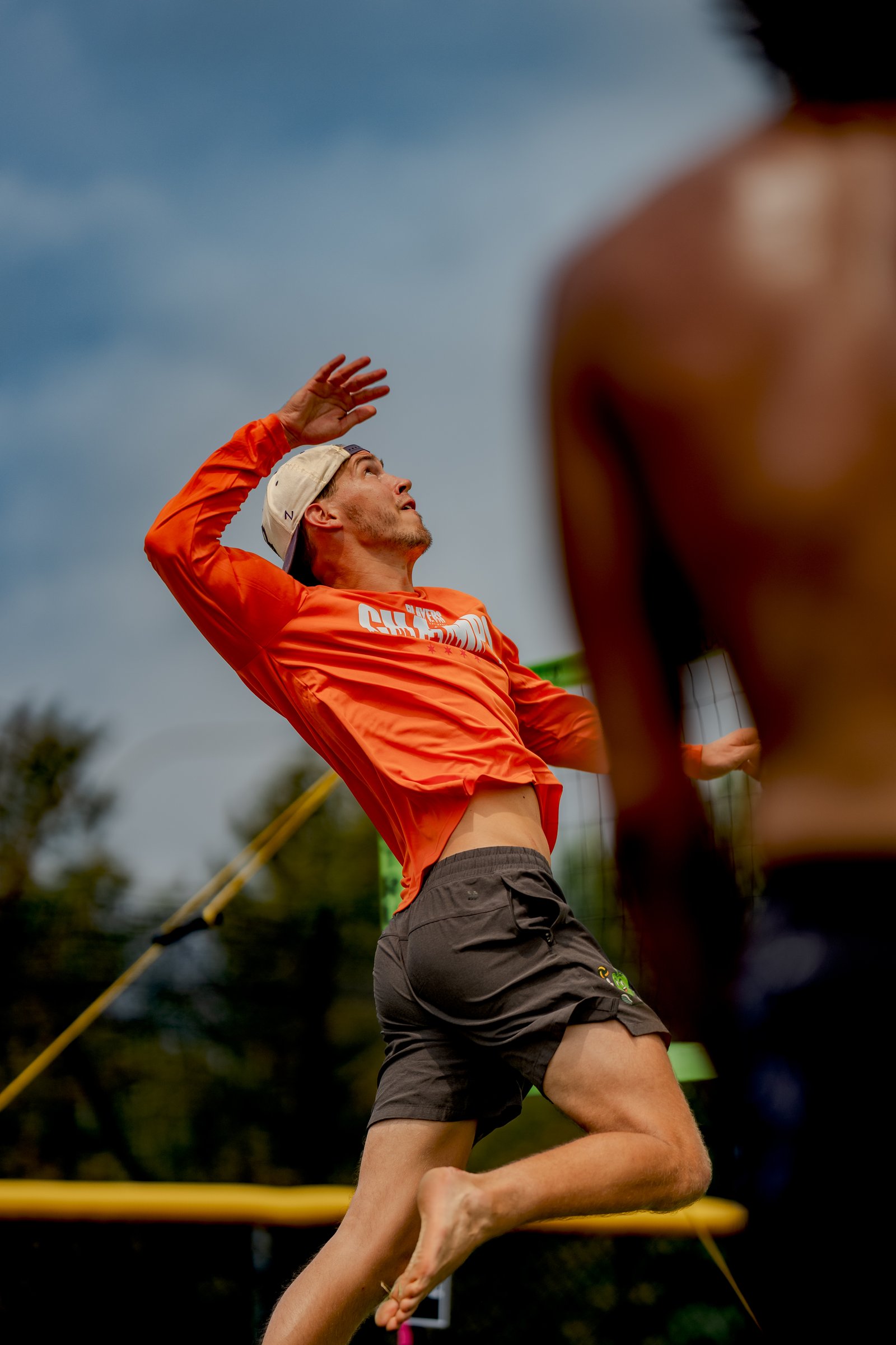 A player in an orange long-sleeve shirt and dark shorts jumps to hit a volleyball, with another player blurred in the foreground.