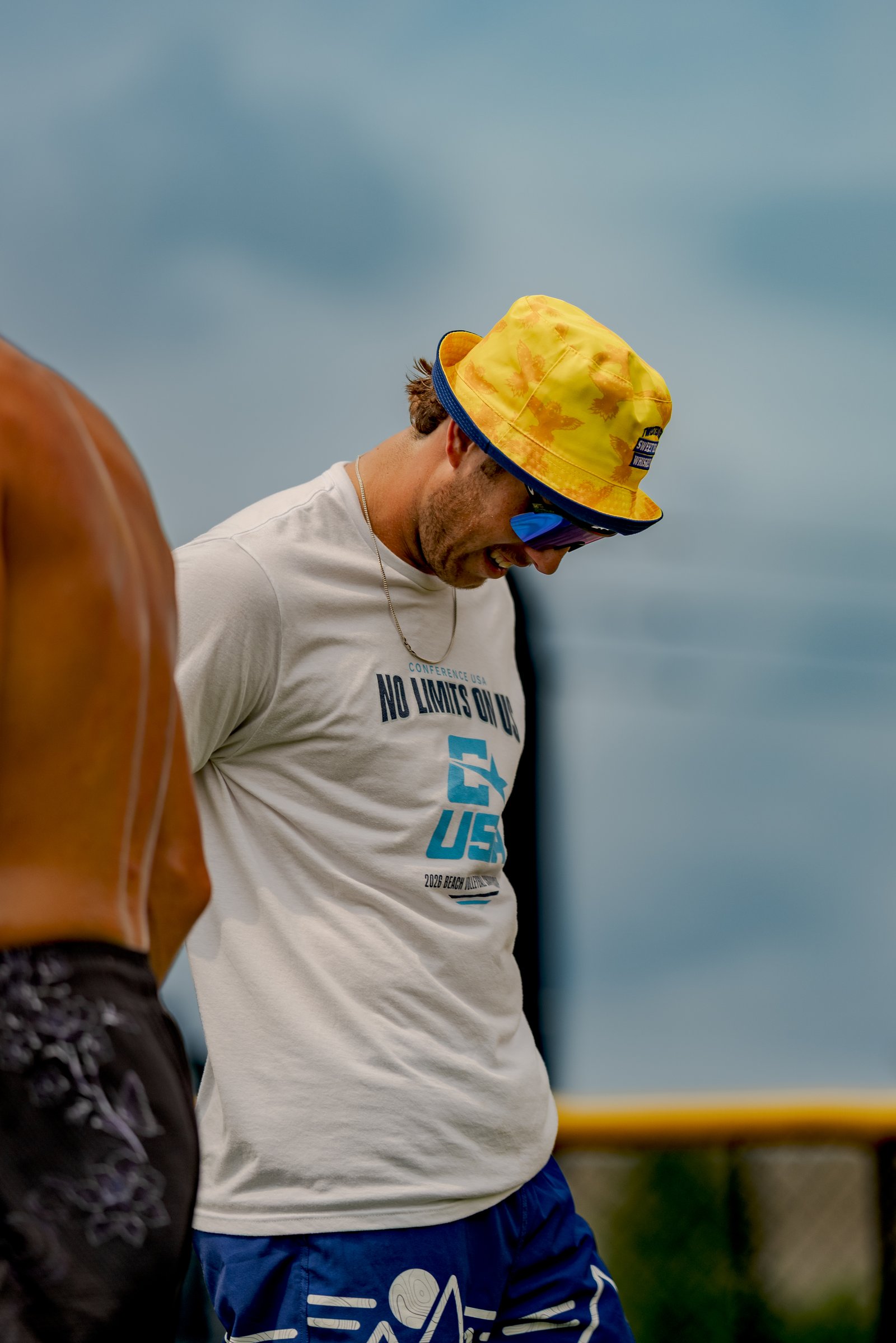 A player in a white shirt and blue shorts with a yellow bucket hat looks down, while another player in black shorts stands nearby.