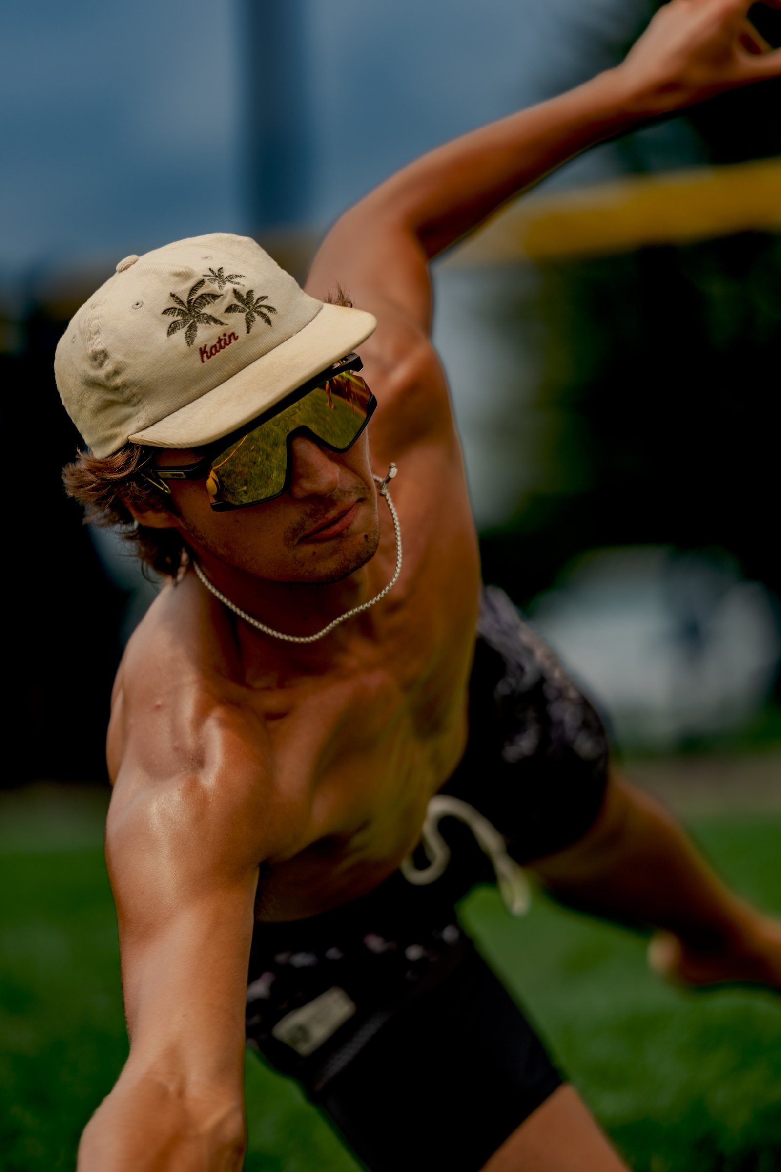 A player in black shorts and a tan cap with palm trees and "Katin" text reaches out on a grassy court.