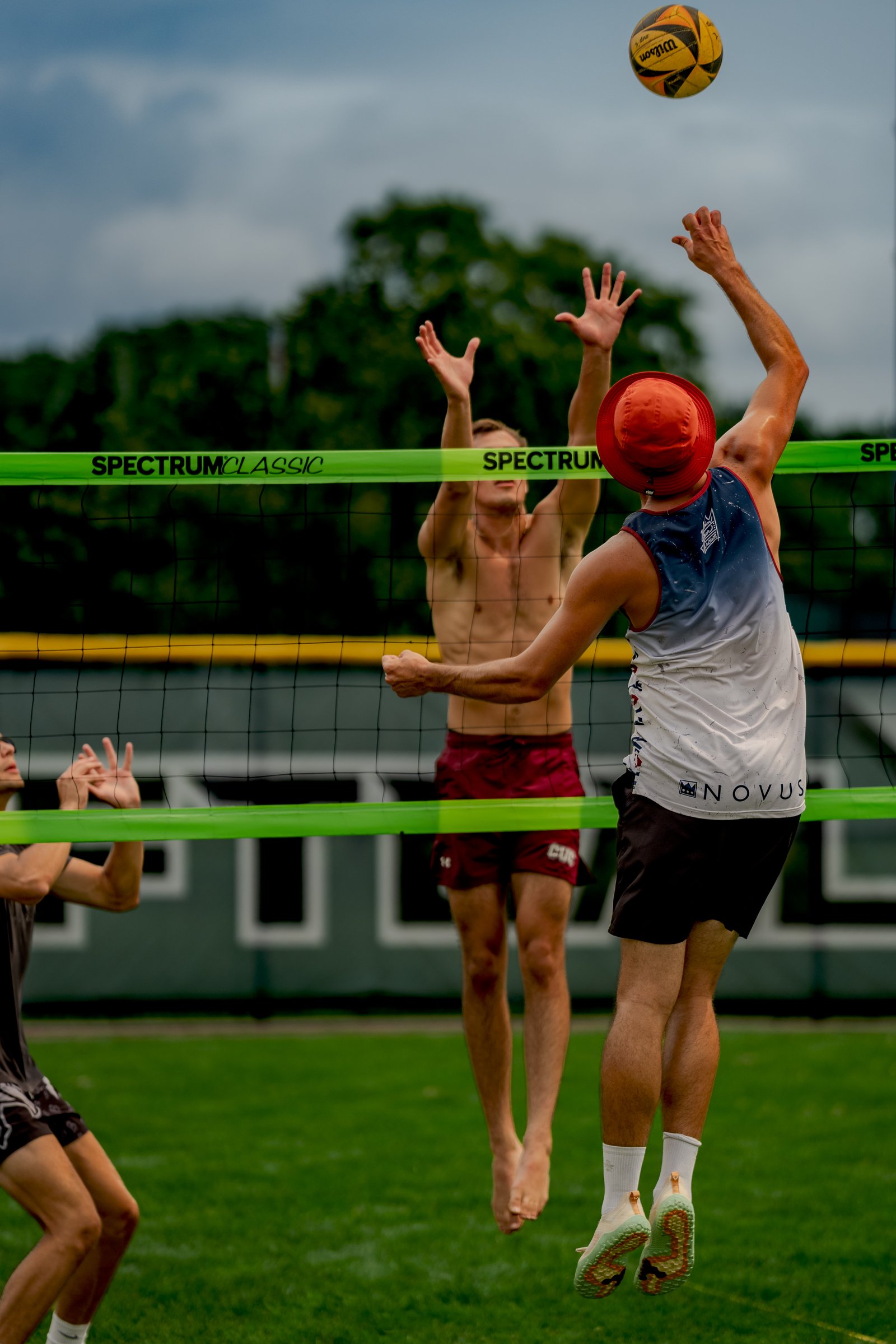 Players in maroon shorts and a white and blue top jump near the net as a volleyball is in the air.