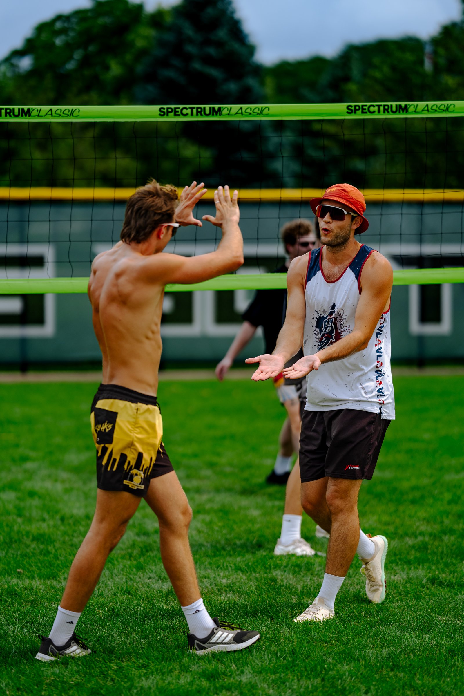 Two players in yellow and black shorts and a white tank top with red trim high-five near a volleyball net on a grassy field.