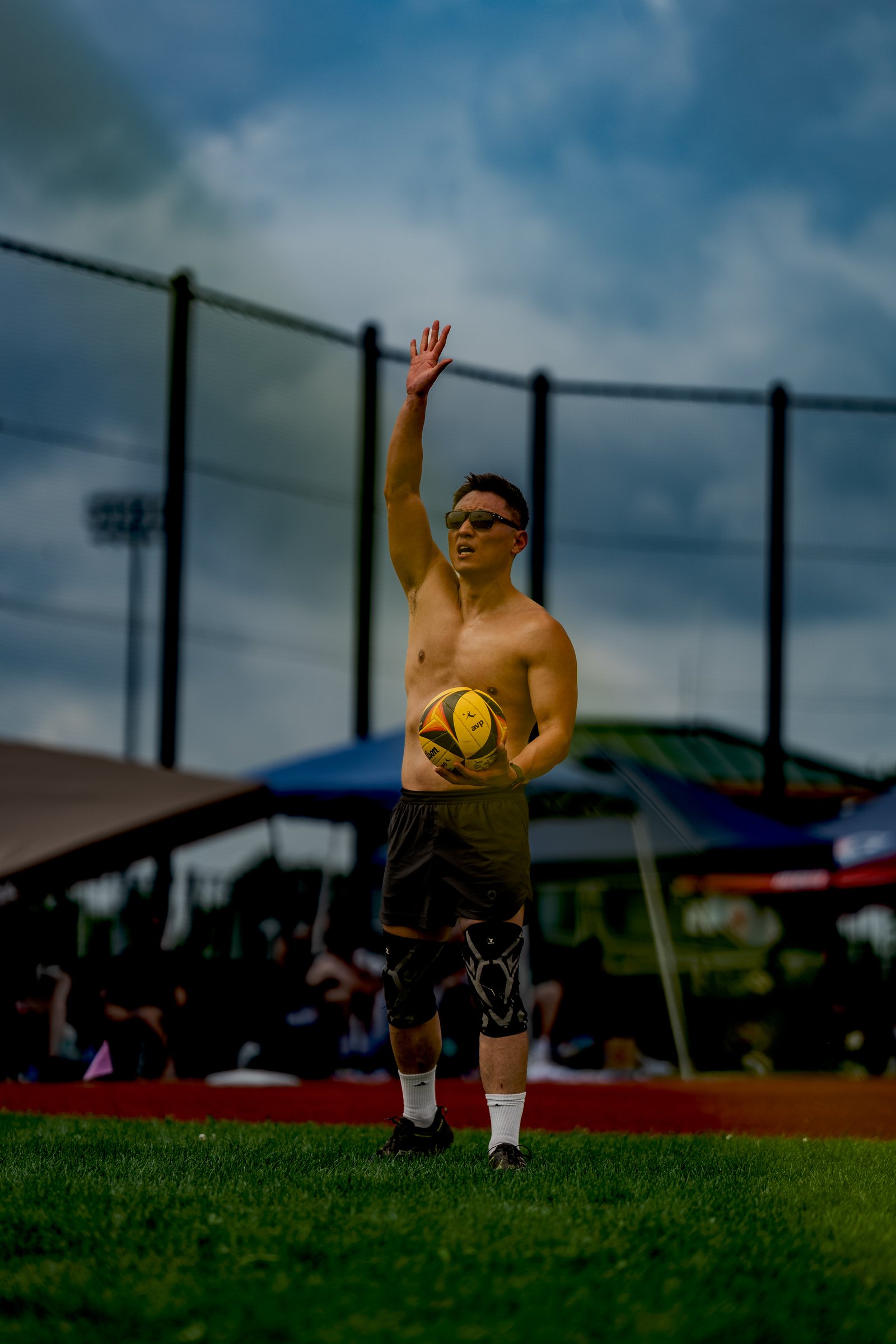 A player in black shorts and knee pads holds a volleyball and raises his hand against a cloudy sky.