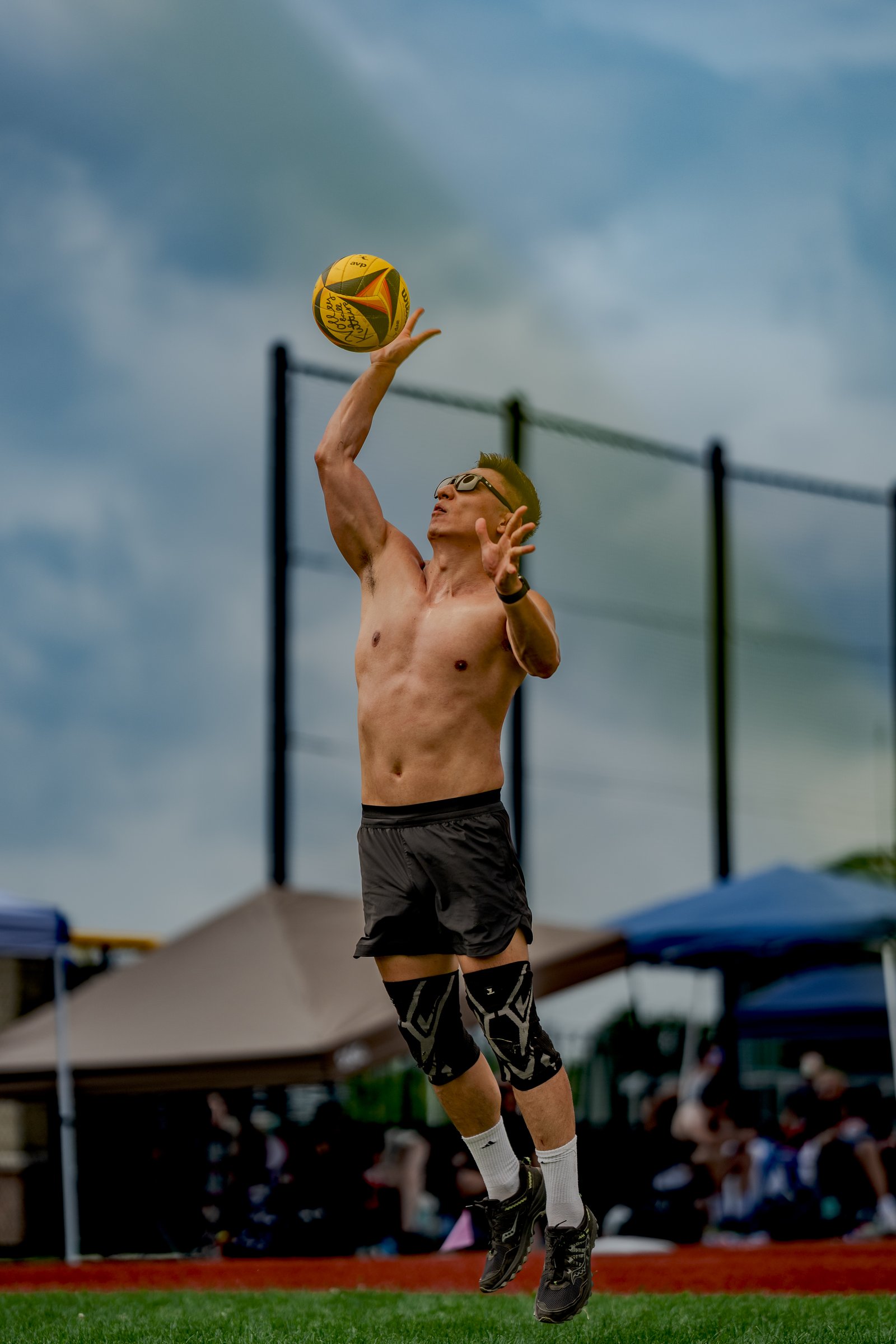 A player in black shorts jumps to serve a volleyball against a cloudy sky with tents and spectators in the background.