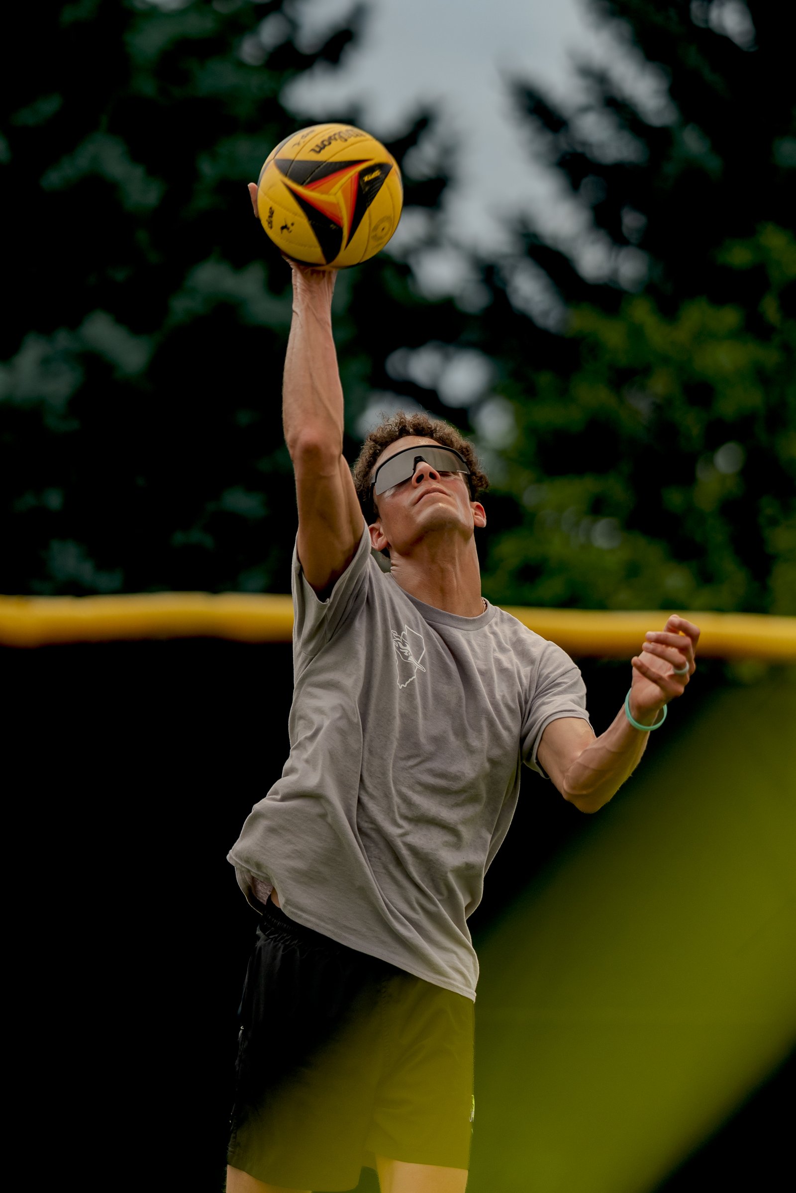 A player in a gray shirt and black shorts reaches up to hit a yellow volleyball.