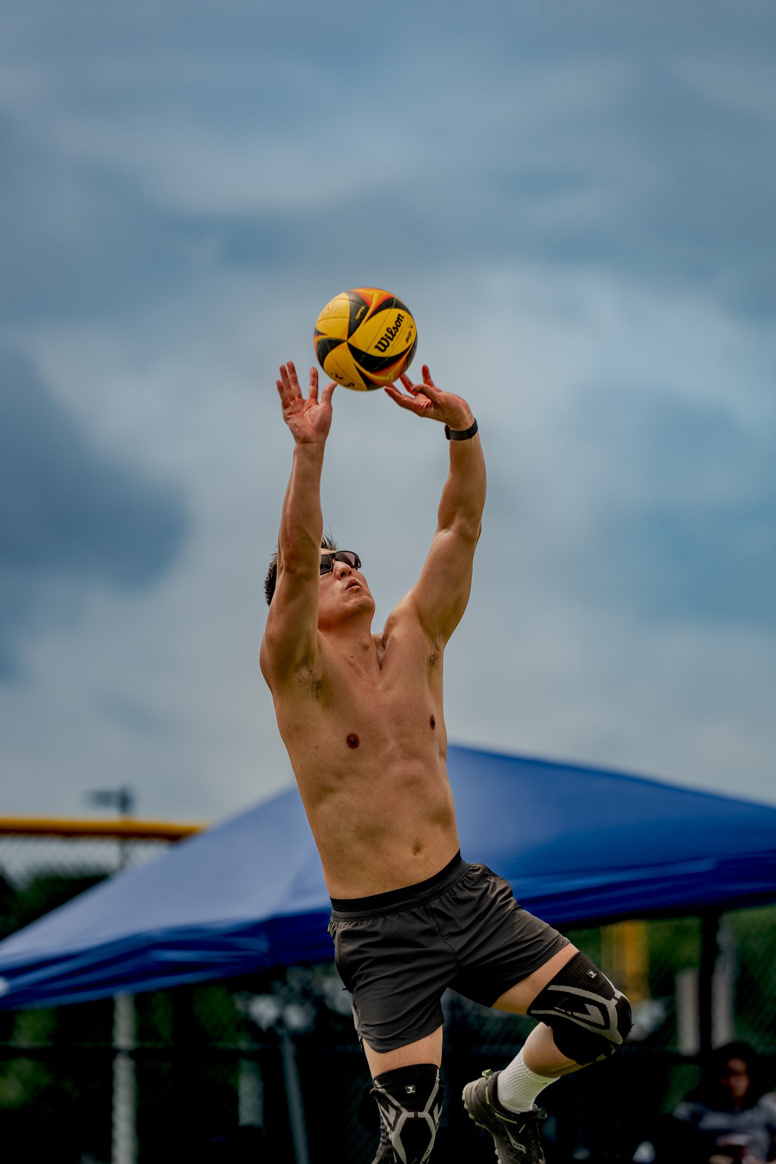 A player in black shorts reaches up to set a volleyball against a cloudy sky.