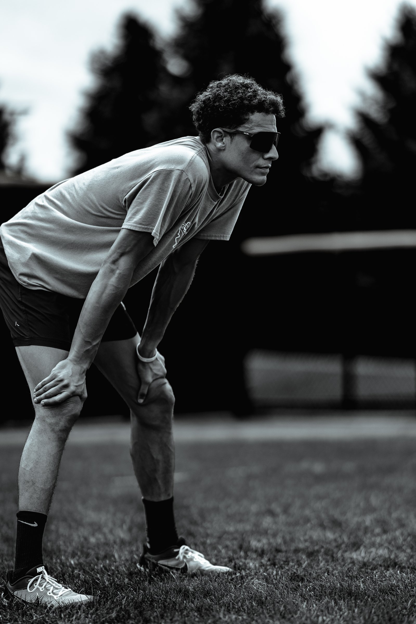 A player in a light-colored shirt and black shorts bends over with hands on knees, wearing sunglasses on a grassy field.